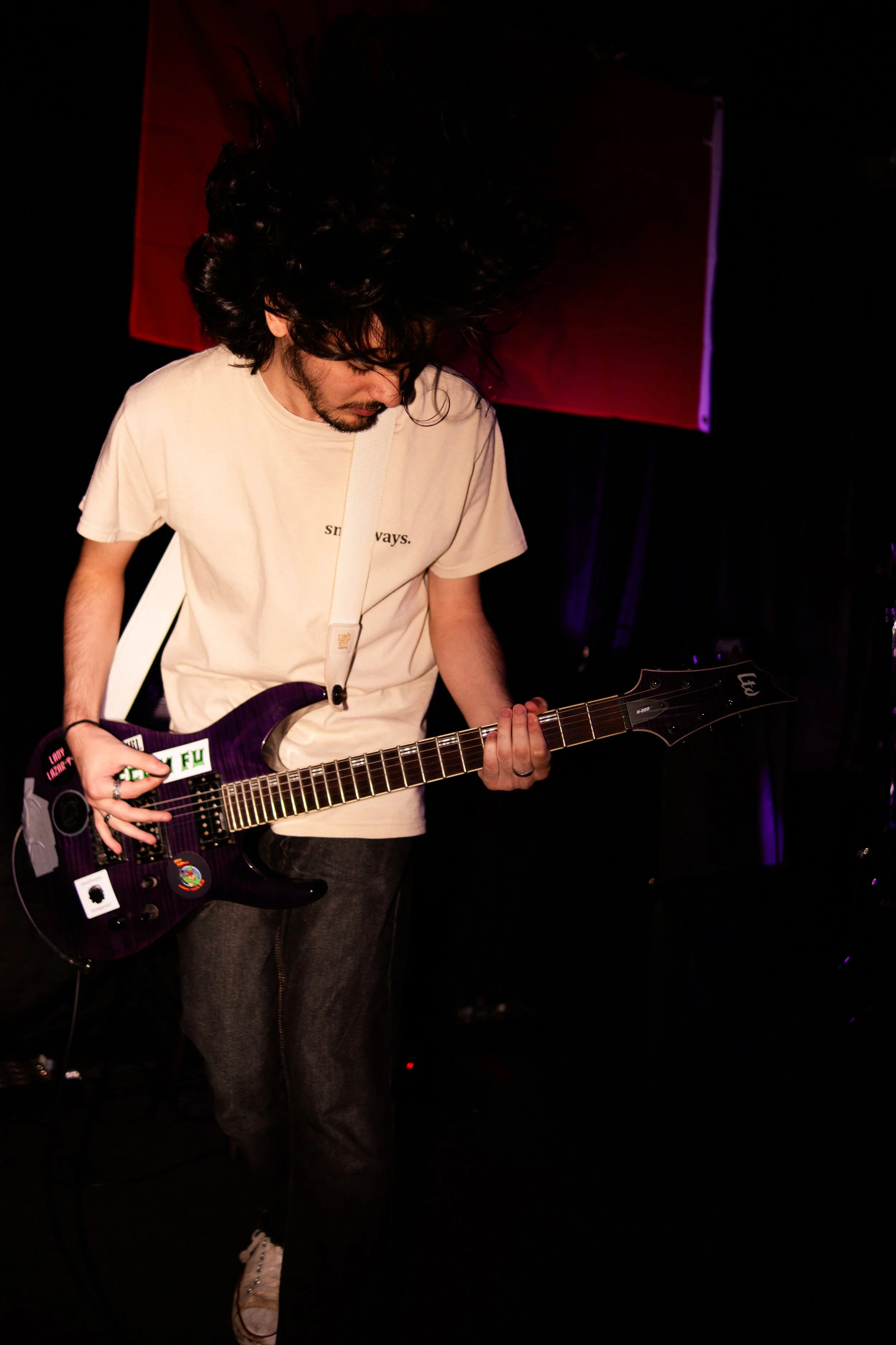 A man with long, curly black hair wearing a beige t-shirt playing an electric guitar on stage in a dark setting with purple lighting.