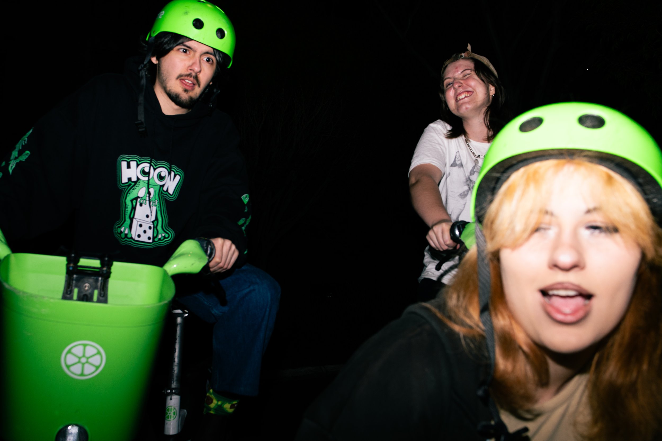 Three young people wearing green helmets riding a green bike or scooter at night.