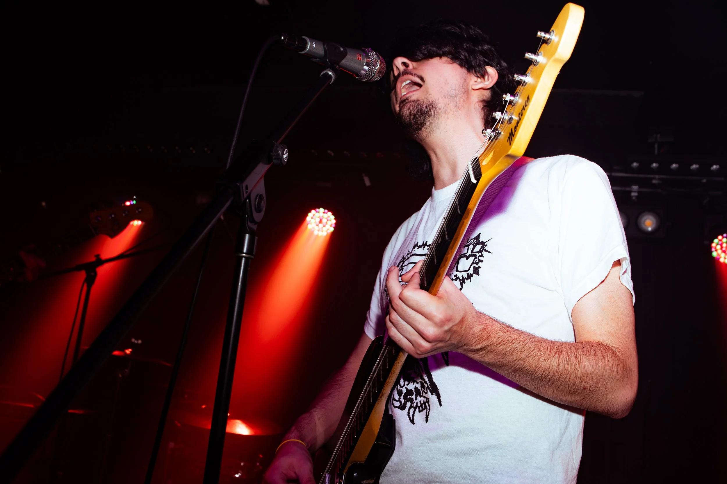 Male musician singing into a microphone while playing an electric guitar on stage with red stage lights.
