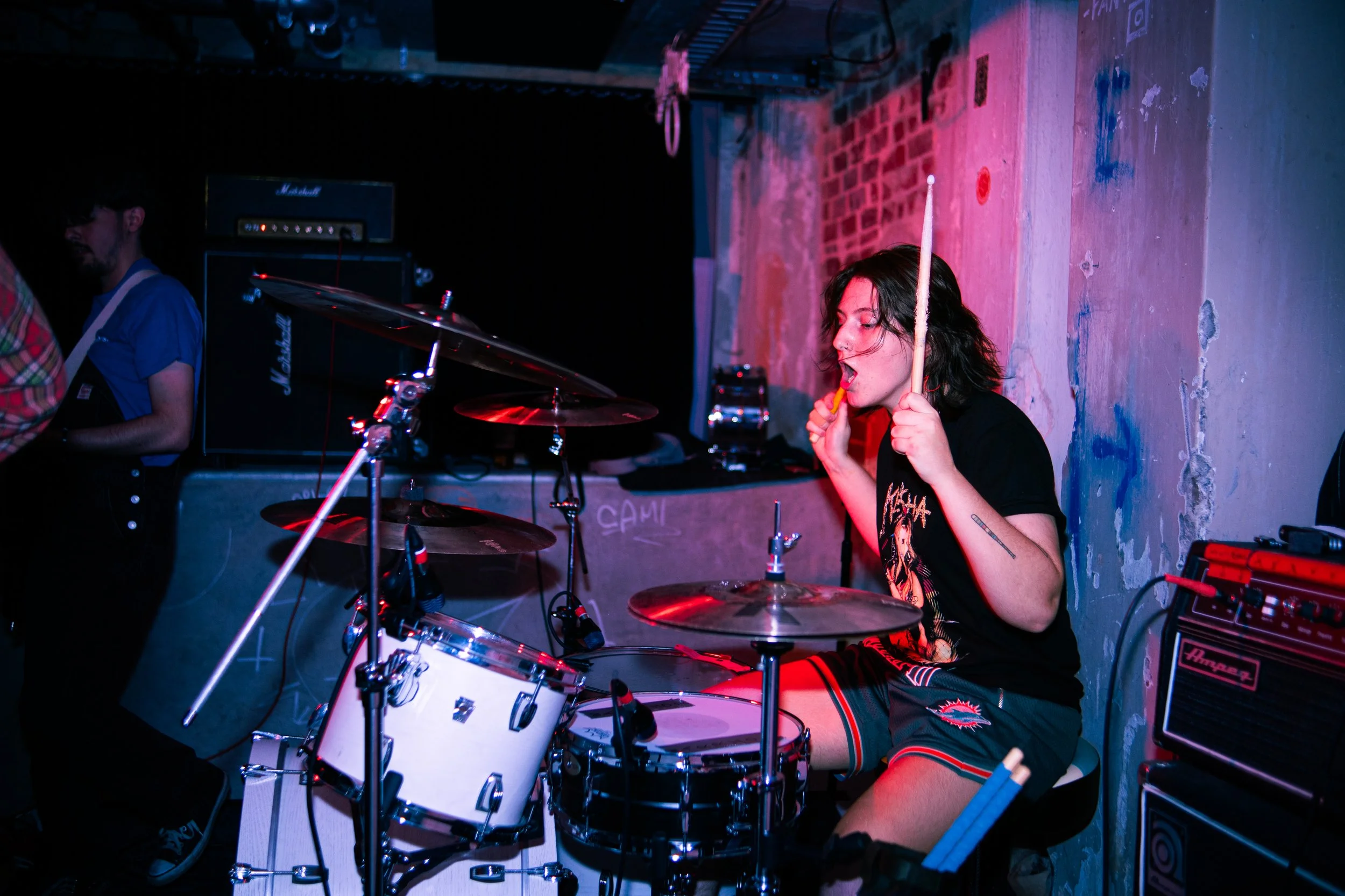 A young woman with dark hair playing drums at a live music venue, surrounded by musical equipment and colorful lighting.