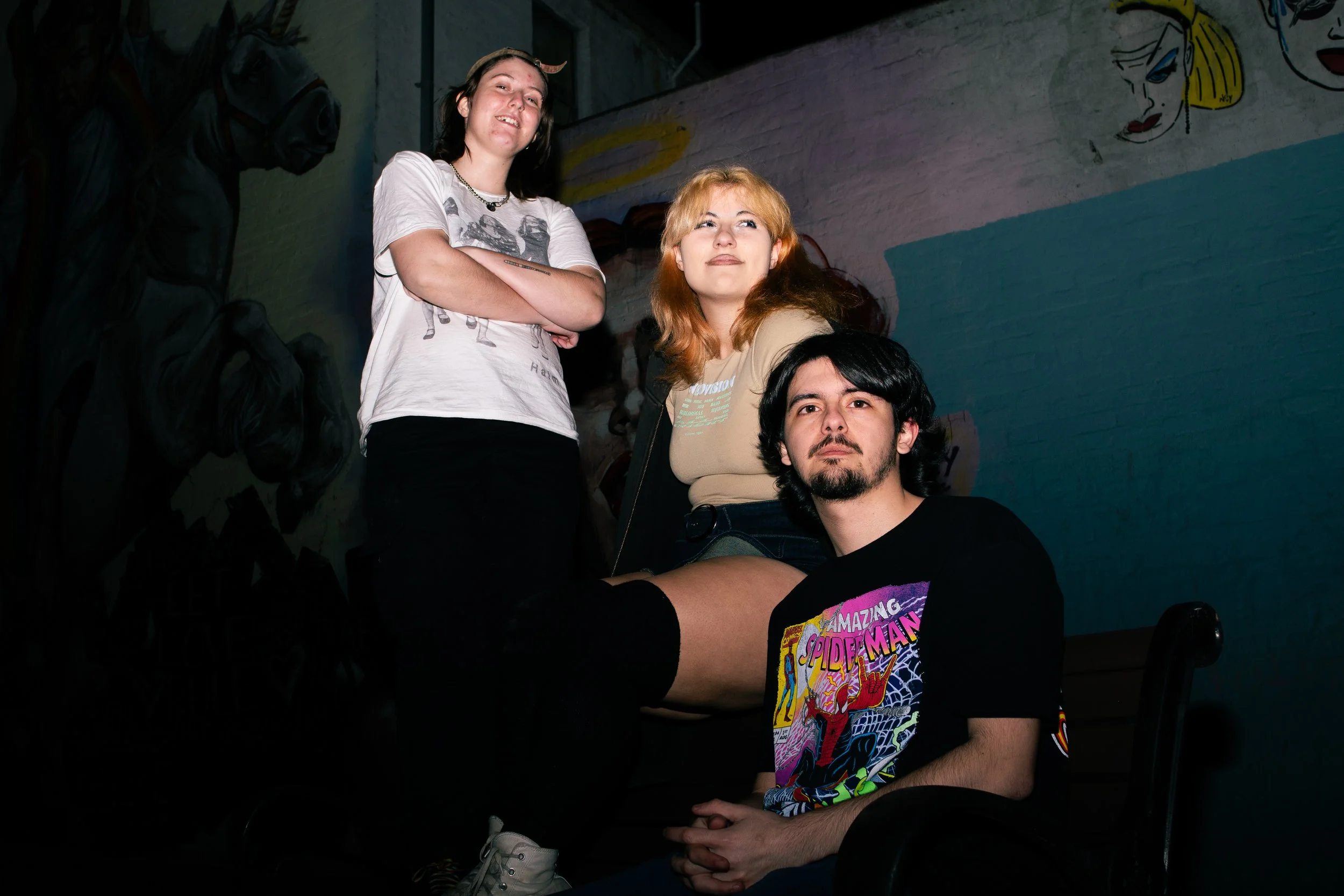 Three young adults posing indoors against a colorful mural on the wall. Two women are standing, one with arms crossed, one with hands behind her back. A young man is sitting on a chair in front, wearing a black T-shirt with a Spider-Man graphic, with