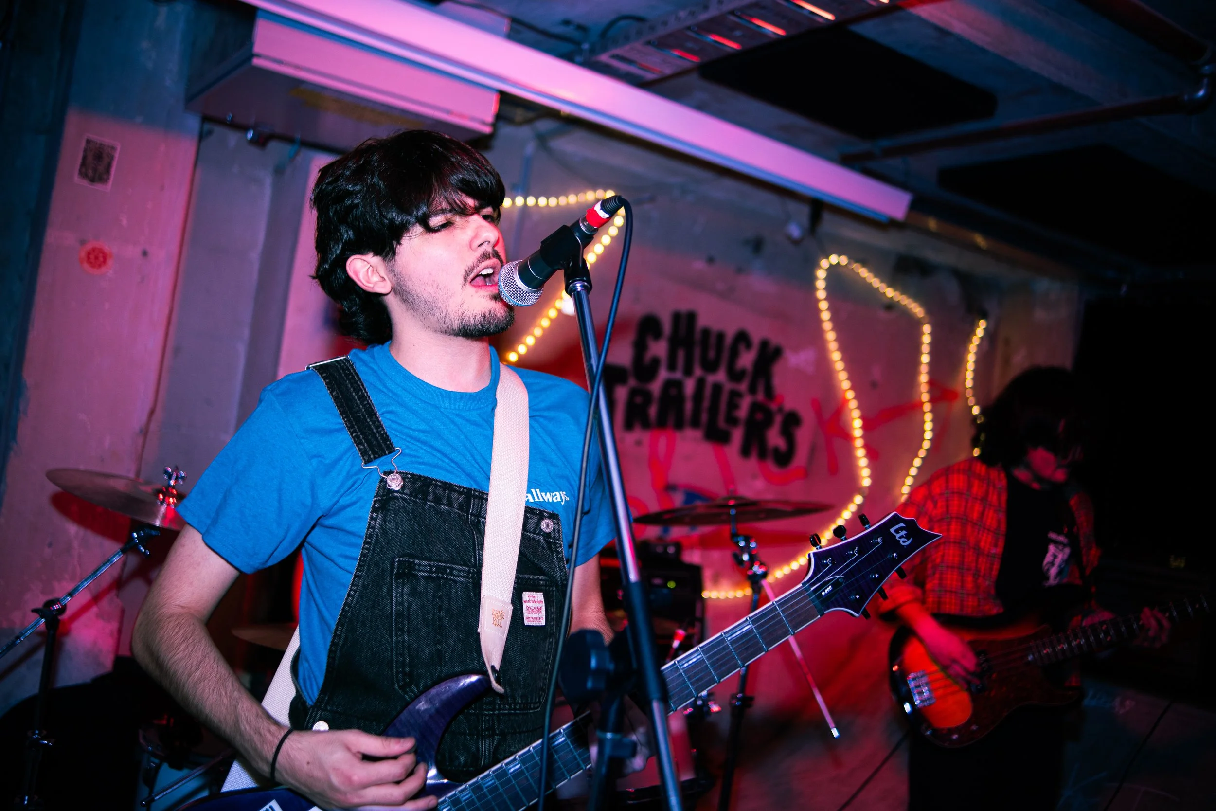 A young man with dark hair, a beard, wearing a blue t-shirt and black overalls, singing into a microphone while playing an electric guitar during a live performance at a venue with a "Chuck Trailers" sign and decorative lights in the background.
