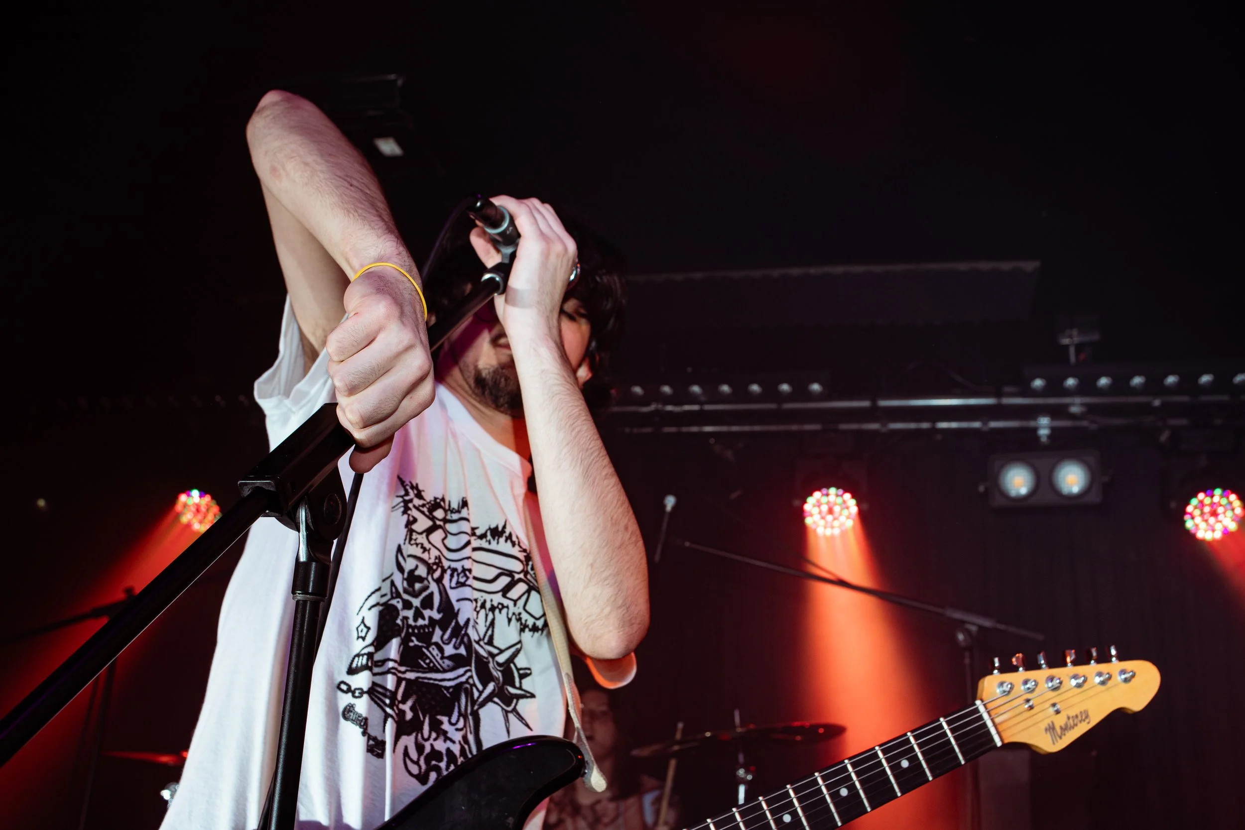 A male musician with dark hair and beard adjusting a microphone on stage with guitar, colorful stage lights in background.