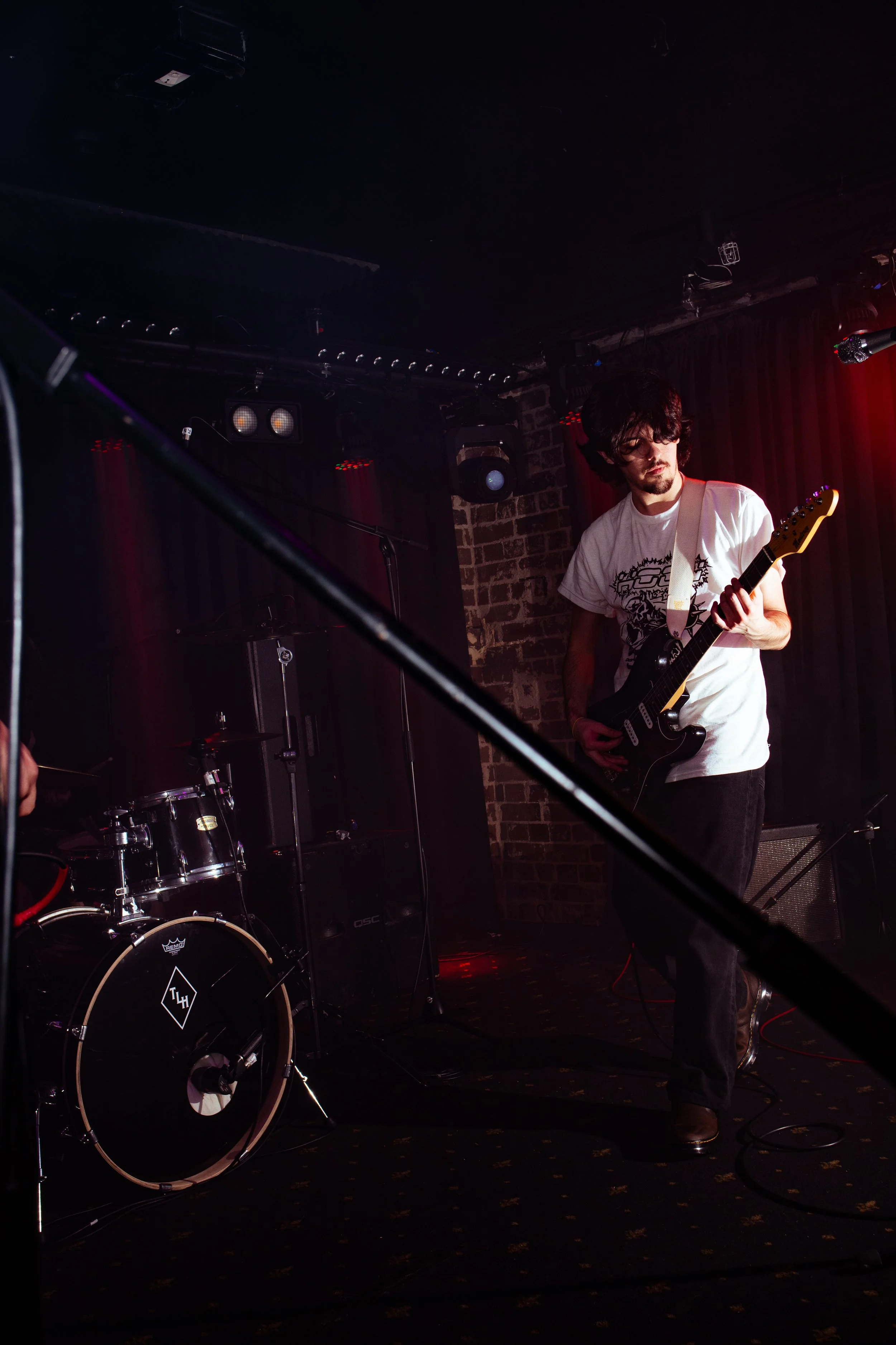 A man playing electric guitar on stage with drums and brick wall background in a dimly lit venue.