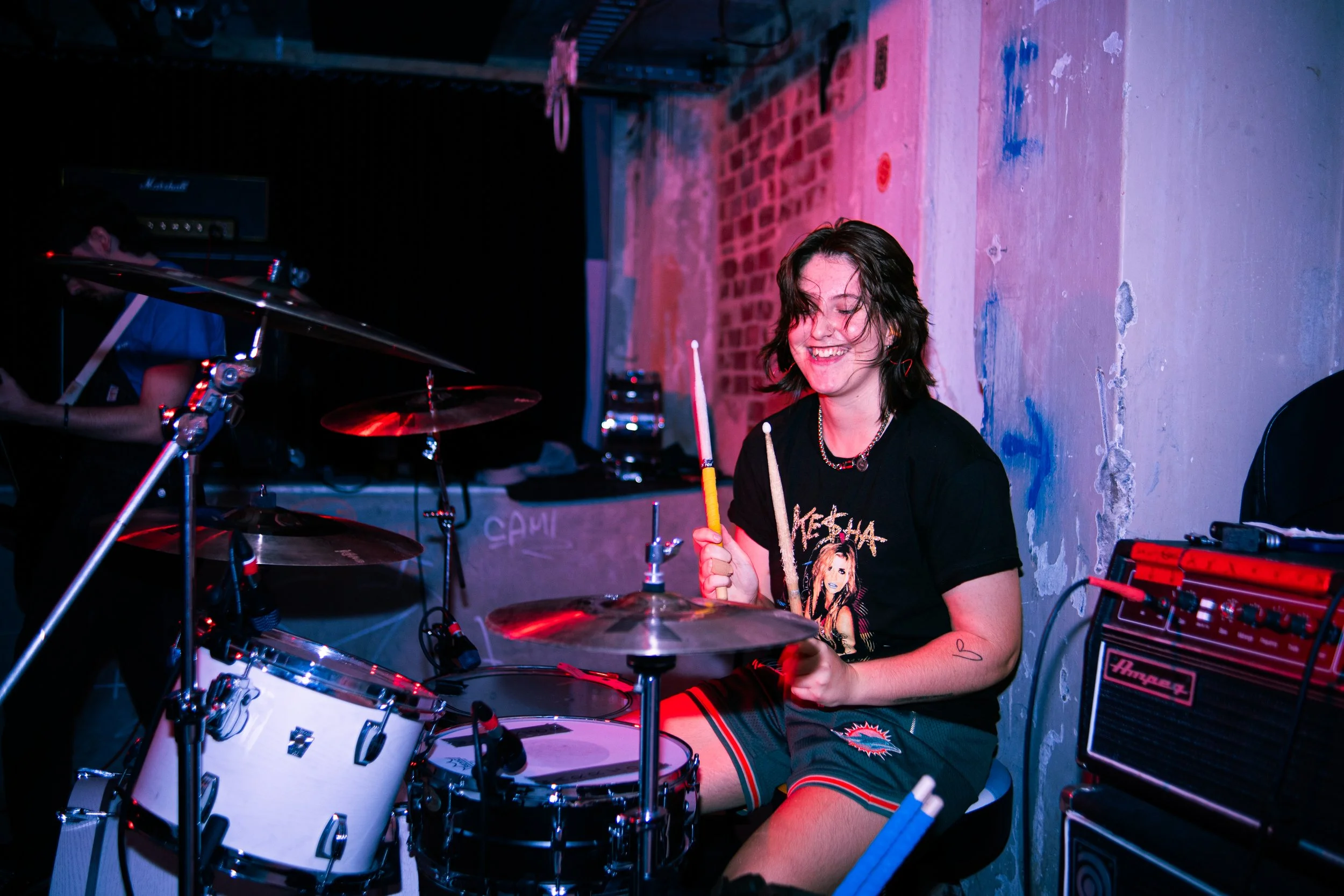 A woman with shoulder-length dark hair playing drums at a music venue, smiling, wearing a black band T-shirt and striped shorts, surrounded by drums and equipment, with a brick and painted wall background.