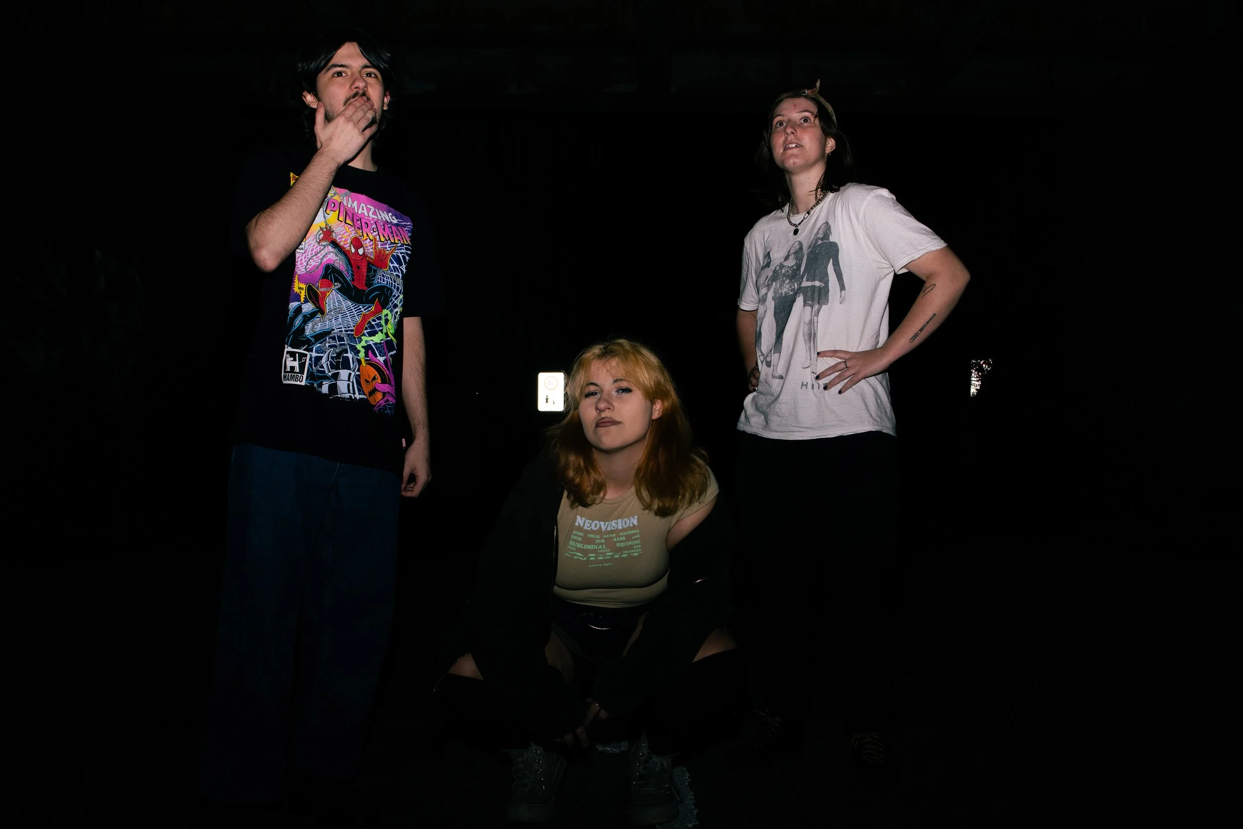 Three young women posing in a dark environment. One is squatting in front, with red hair and a graphic green t-shirt. The other two are standing, one with long dark hair and a white t-shirt, the other with short dark hair and a black graphic t-shirt.