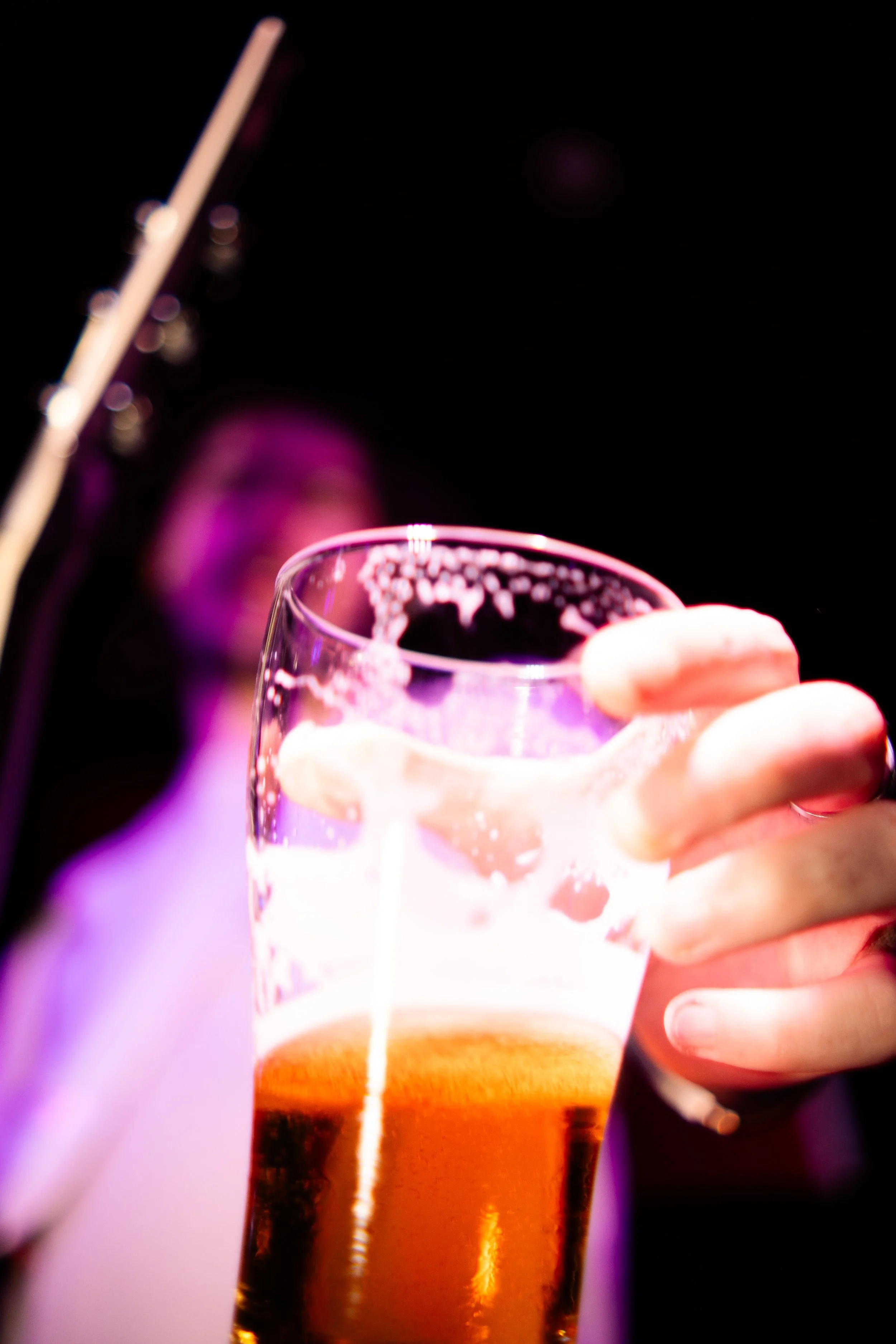 Person holding a glass of beer in a bar or nightclub setting, with a blurred background and purple lighting.