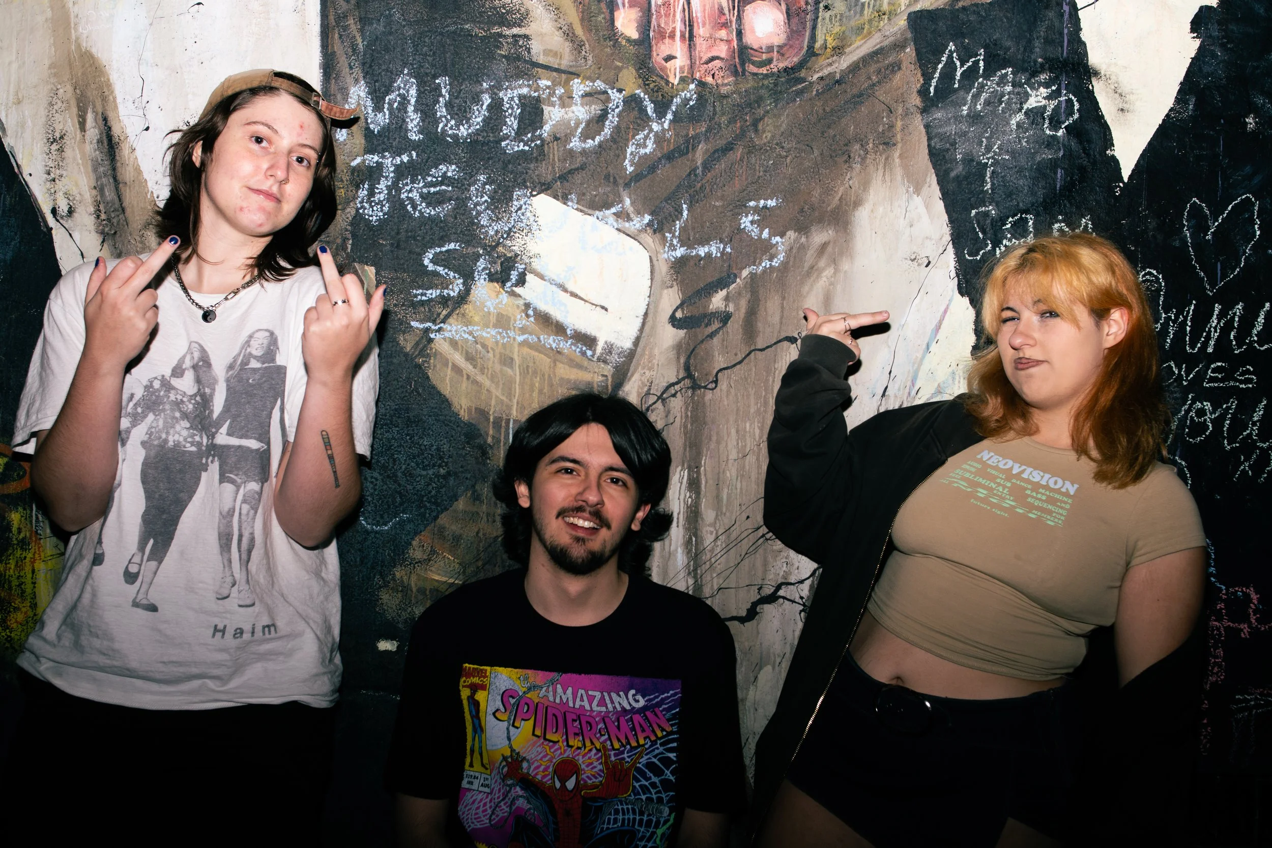 Three young people in front of a graffiti-style mural, two women and one man, all making hand gestures.