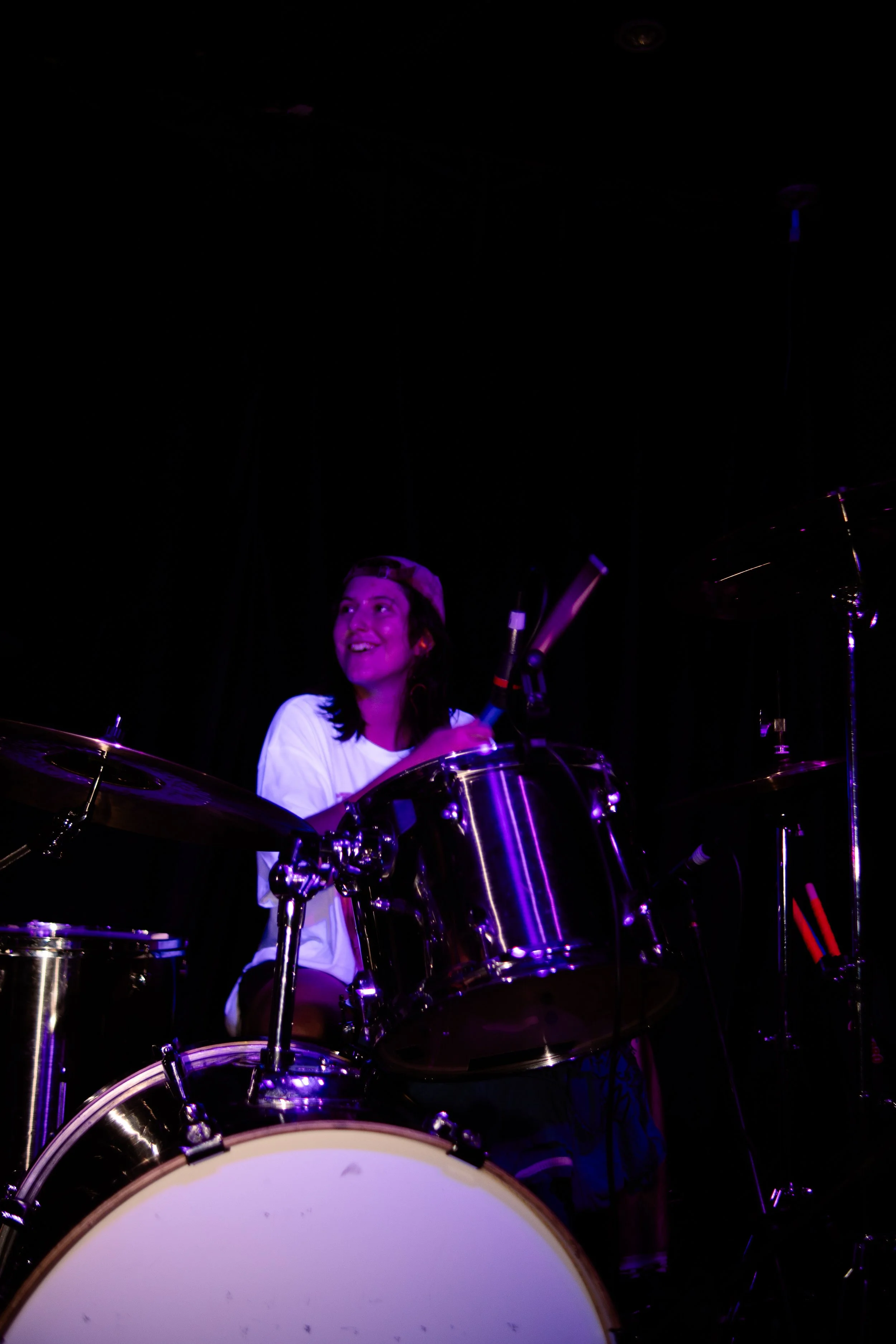 A young woman playing drums on stage, smiling, wearing a backward cap and a white t-shirt, illuminated by purple stage lighting.