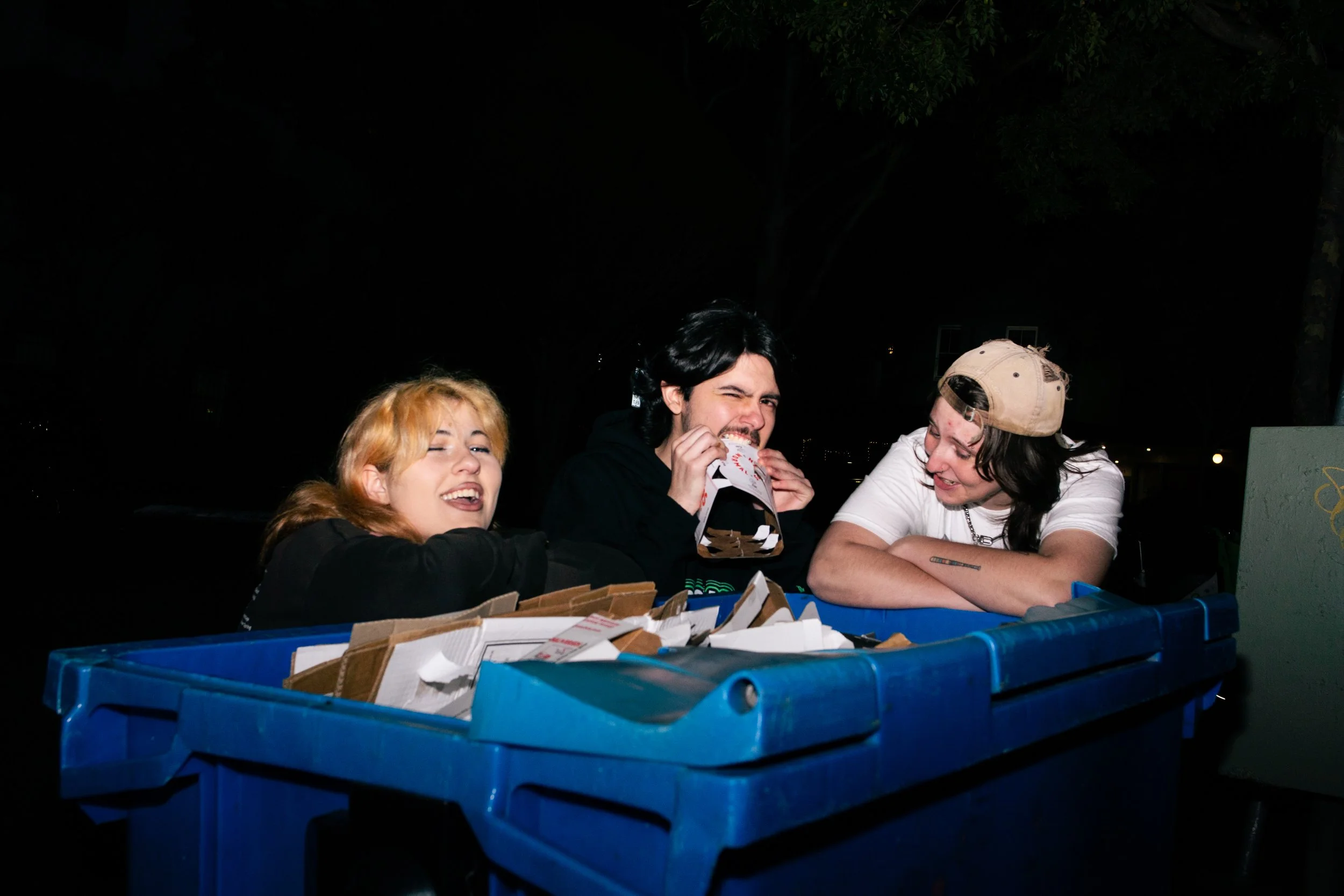 Three young people sitting around a blue recycling bin filled with paper food packaging, at night. They are smiling and laughing, with one person biting into a cardboard pizza box, in a park setting.