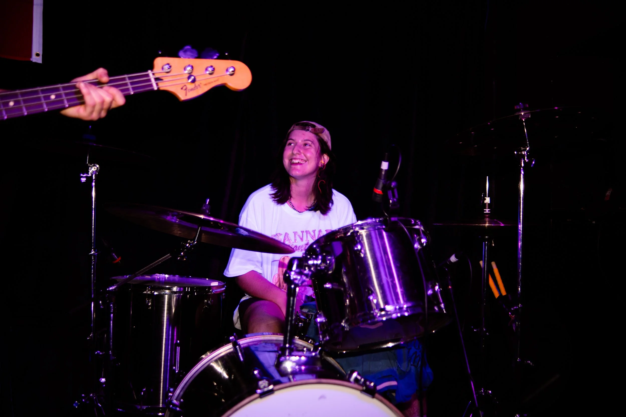 A young woman sitting behind a drum set, smiling and looking up, with purple stage lighting. A person's hand playing a guitar is partially visible on the left side of the image.