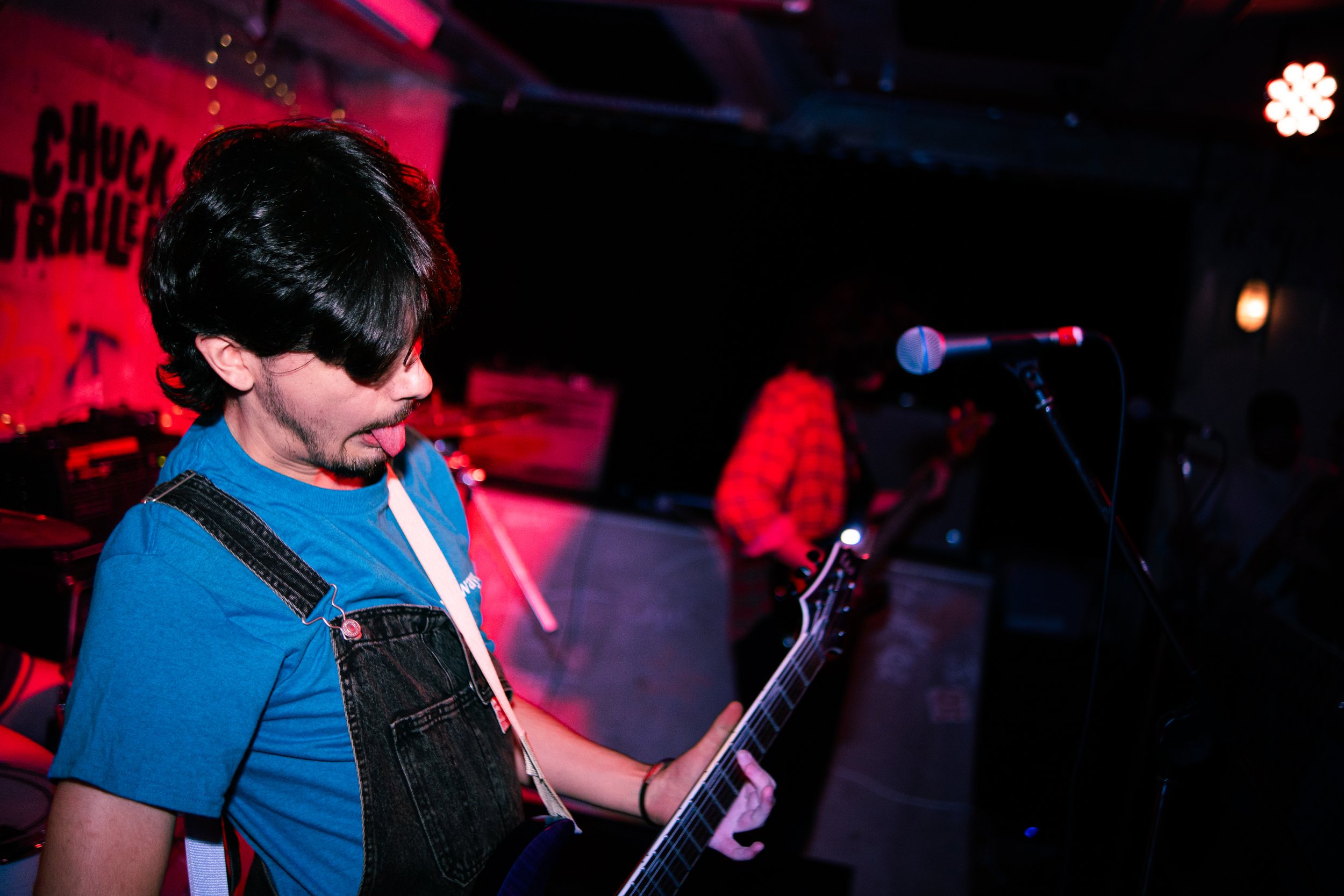 Musician with dark hair, blue t-shirt, playing an electric guitar on stage with a red backdrop and a person in a red checkered shirt in the background.