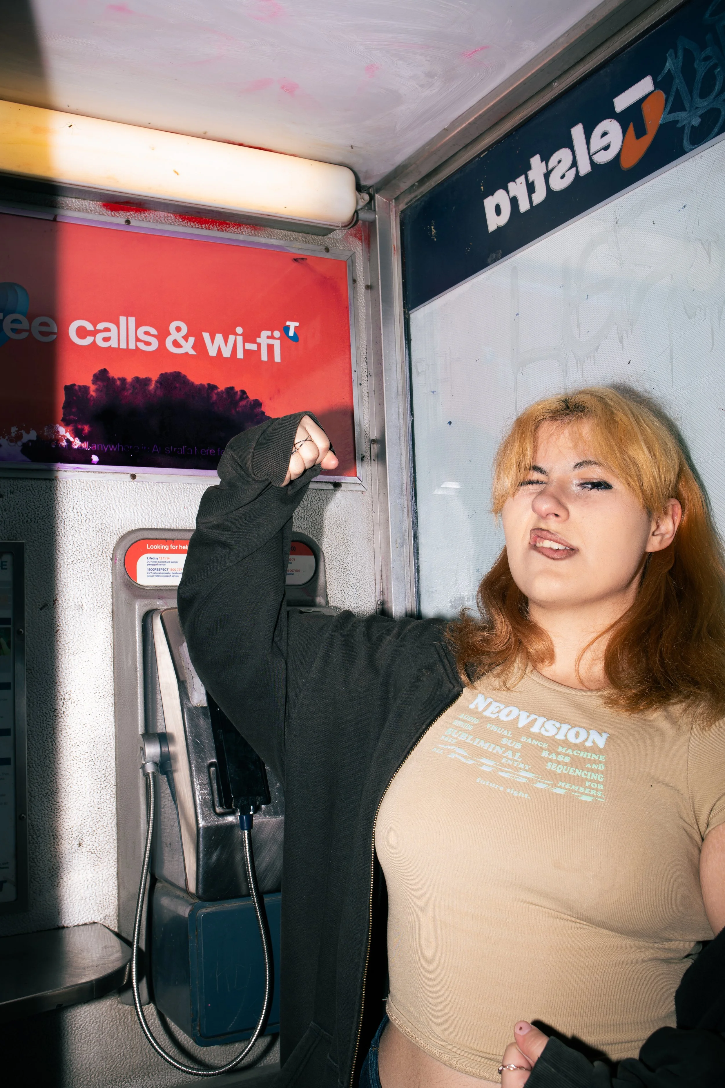 A young woman with orange hair making a fist in front of a payphone, inside a booth with signage about free calls and Wi-Fi.