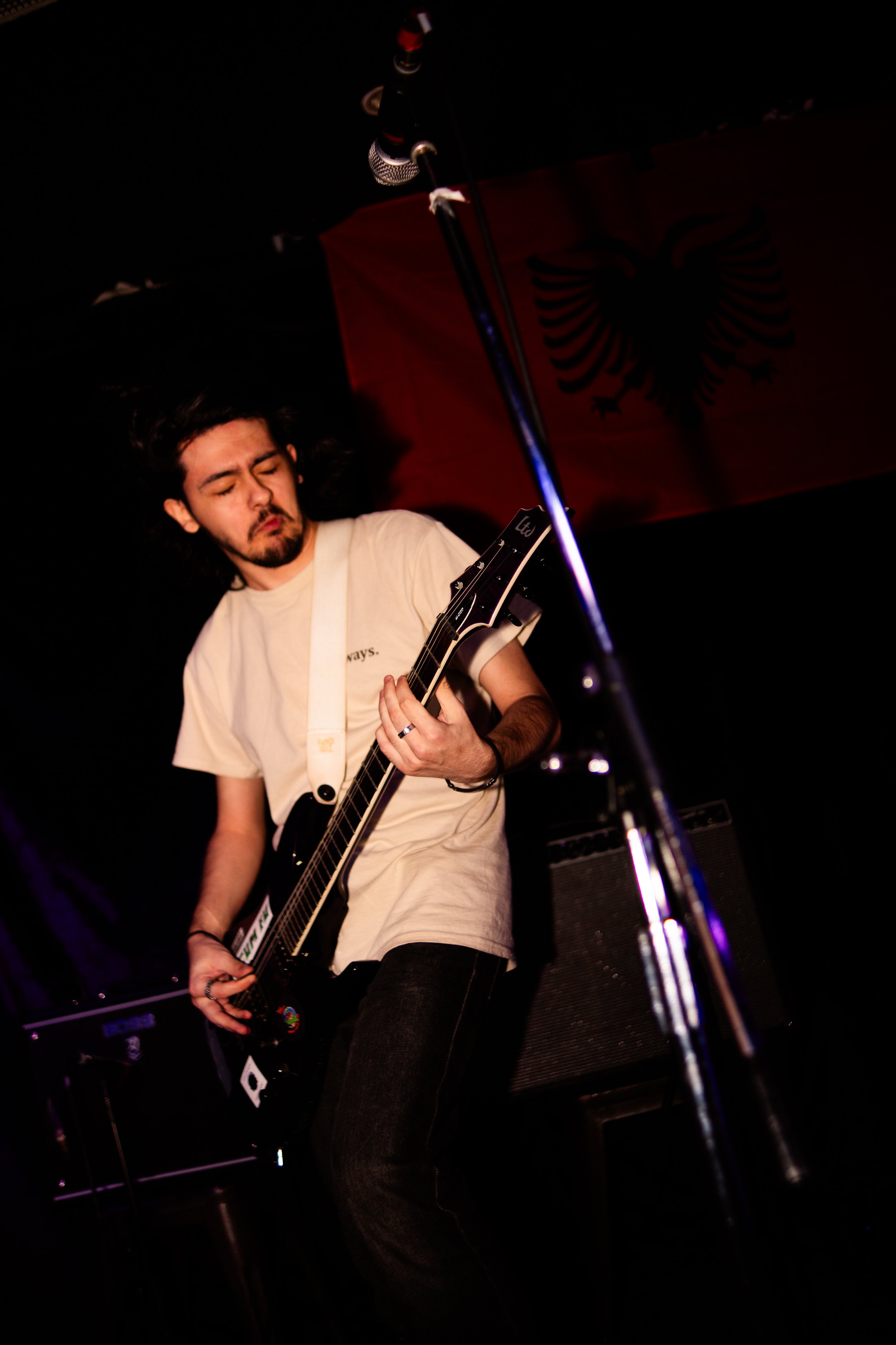 A man with long dark hair playing an electric guitar on stage, with a red and black flag in the background.