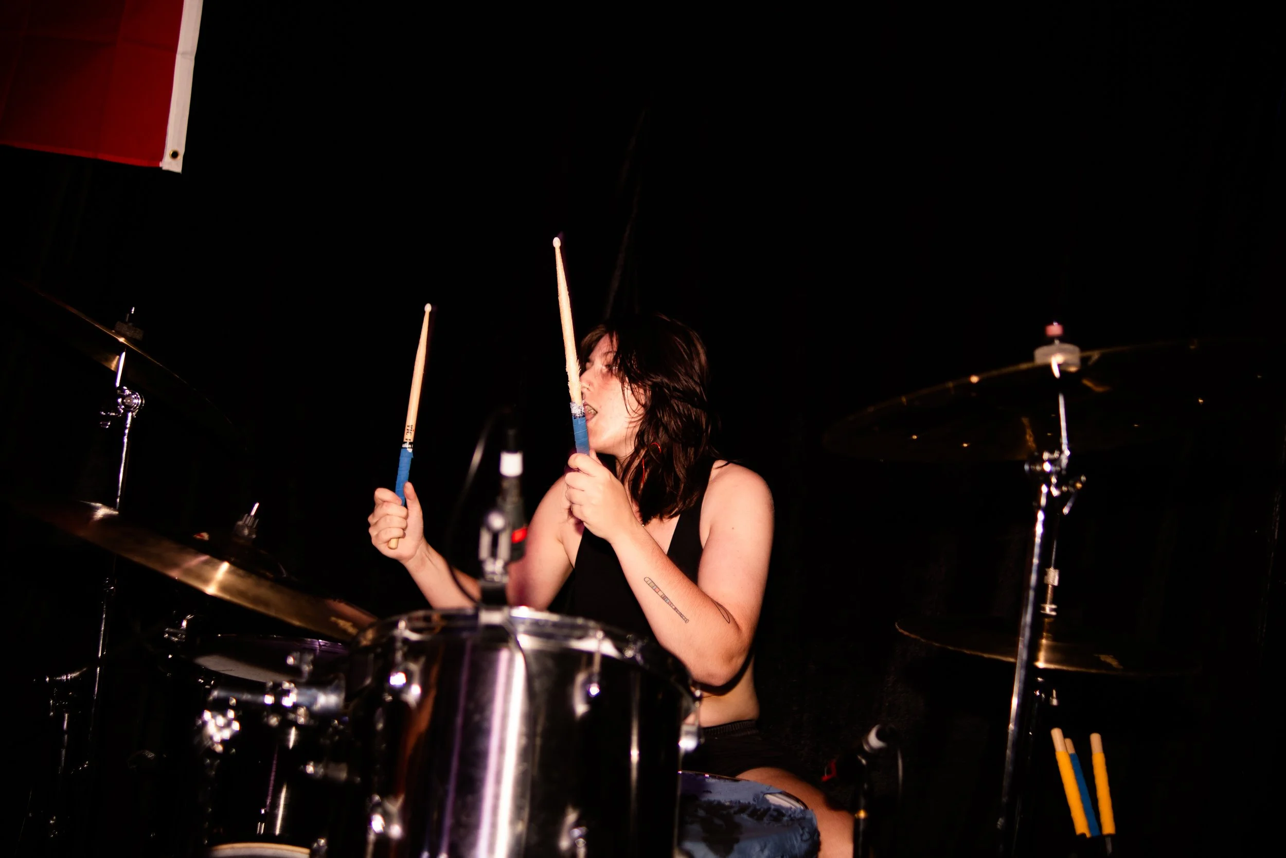 A woman playing drums on stage, holding drumsticks near her face, with a dark background.