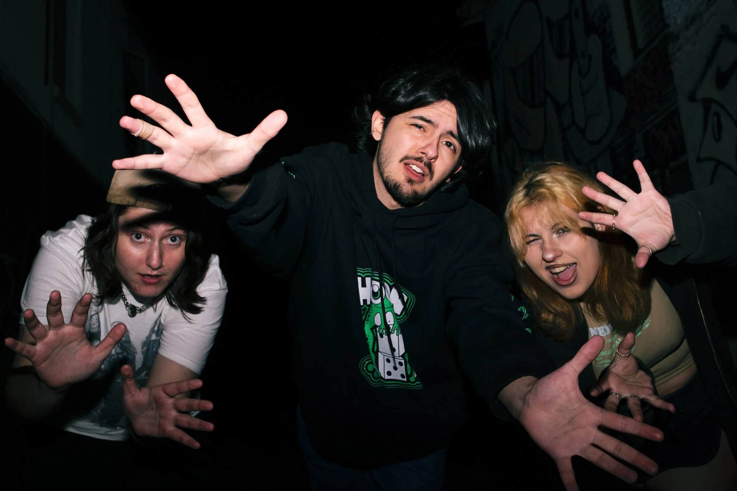 Group of three young adults making exaggerated expressive faces and hand gestures in a dark setting, one with black hair, one with blond hair, and one with dark brown hair.