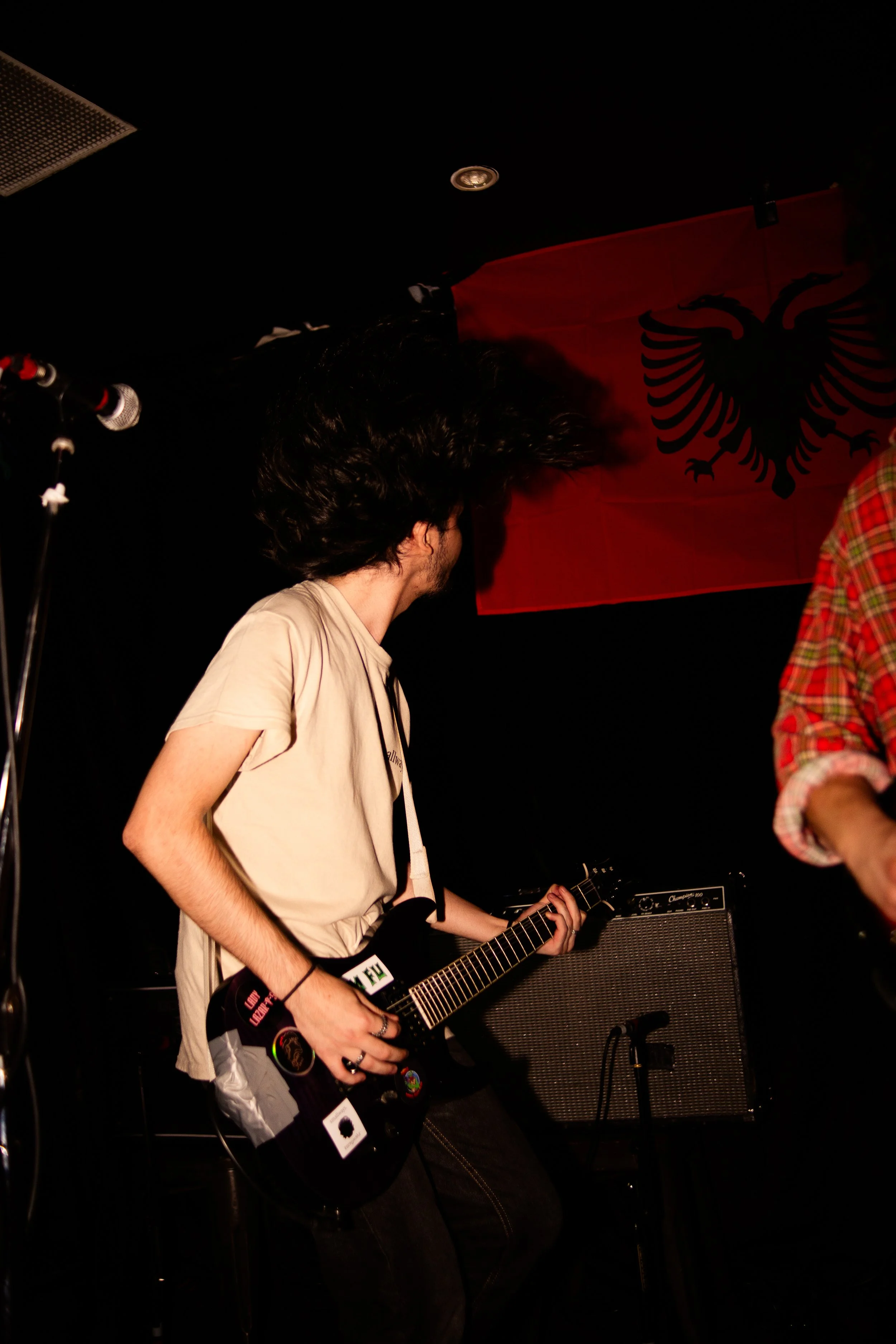 A person playing an electric guitar on stage with a red flag featuring a black double-headed eagle in the background.