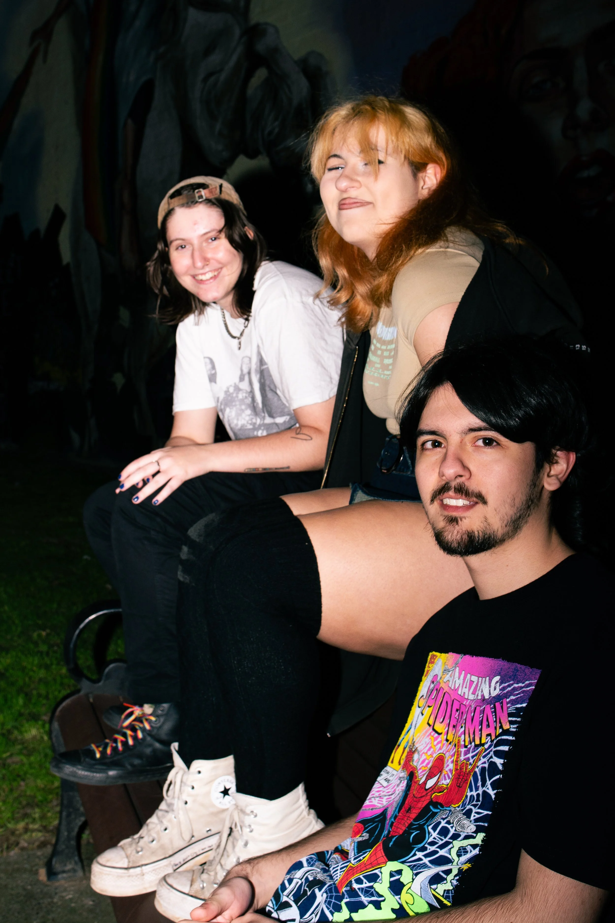 Three friends sitting on a park bench at night, smiling at the camera, with a dark mural background.
