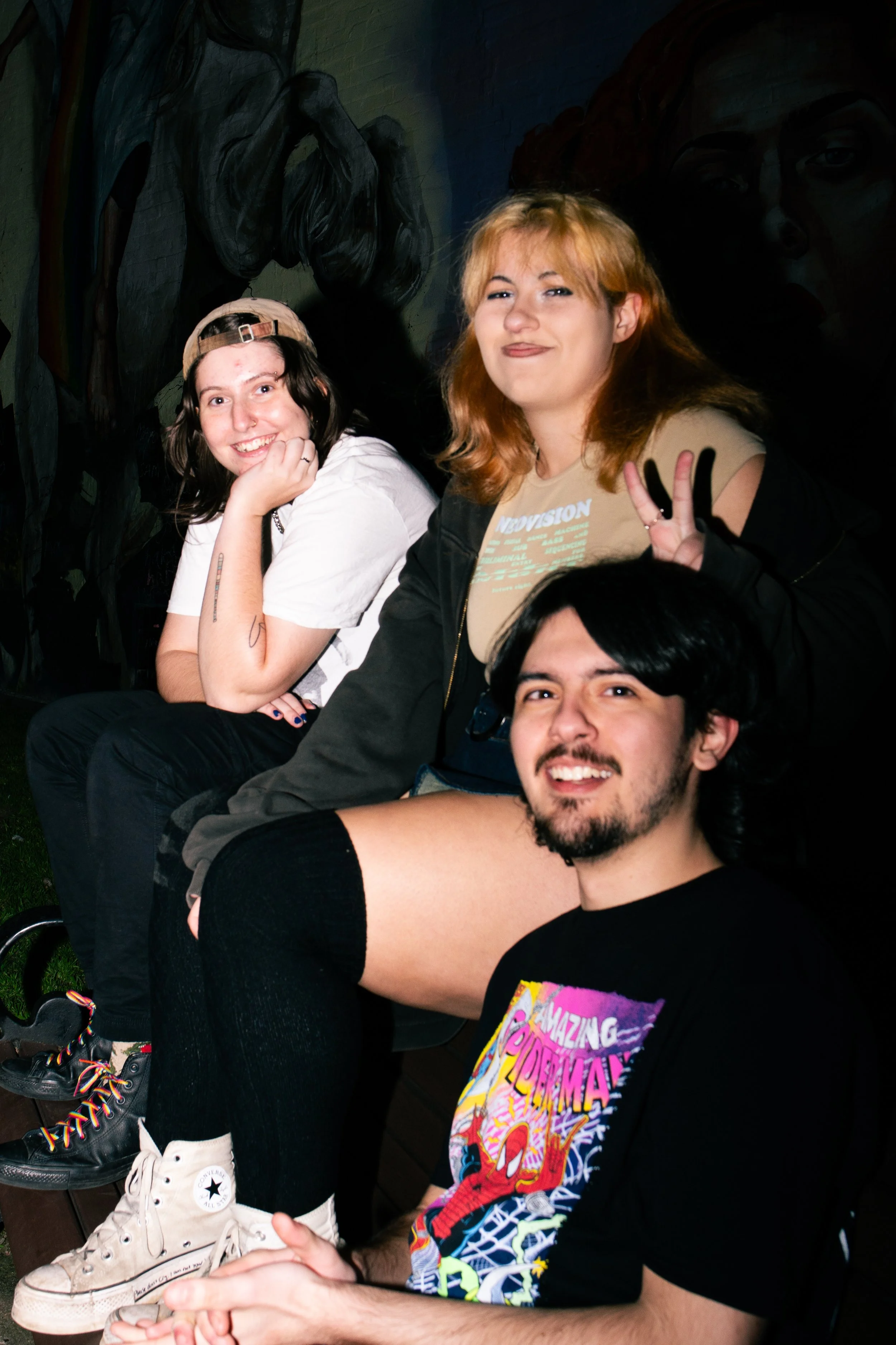 Three friends sitting outside at night, smiling and making gestures. A girl in a white shirt and a beige cap, a girl in a beige t-shirt with a black jacket, and a guy in a black Spider-Man t-shirt sitting on the ground with his leg up.