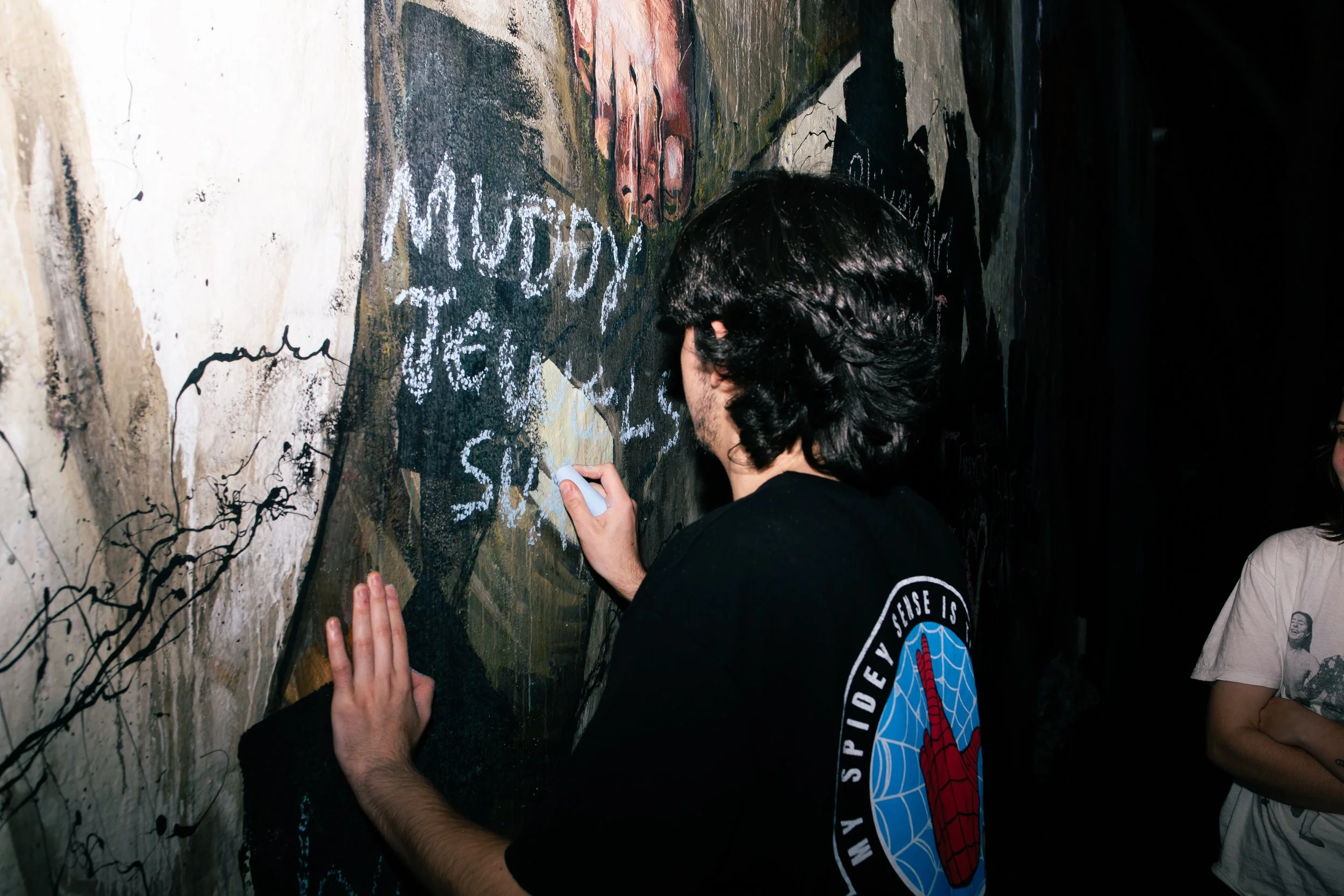 A person with black hair writing on a dark wall with white chalk, with a child standing nearby watching, in a dimly lit environment.