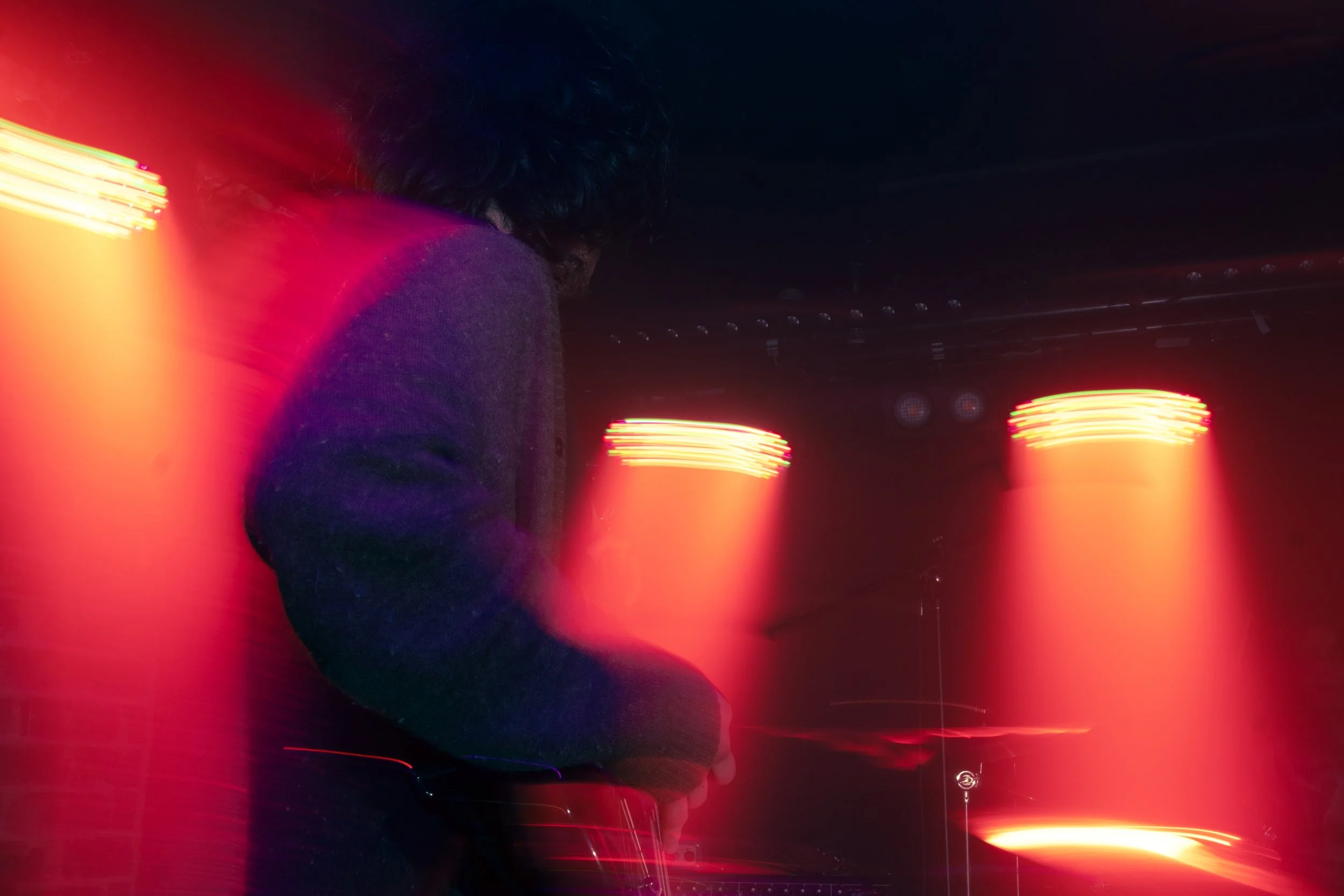 A person with dark, curly hair and a purple sweater is performing as a DJ in a dark nightclub, surrounded by colorful red, orange, and pink stage lights.