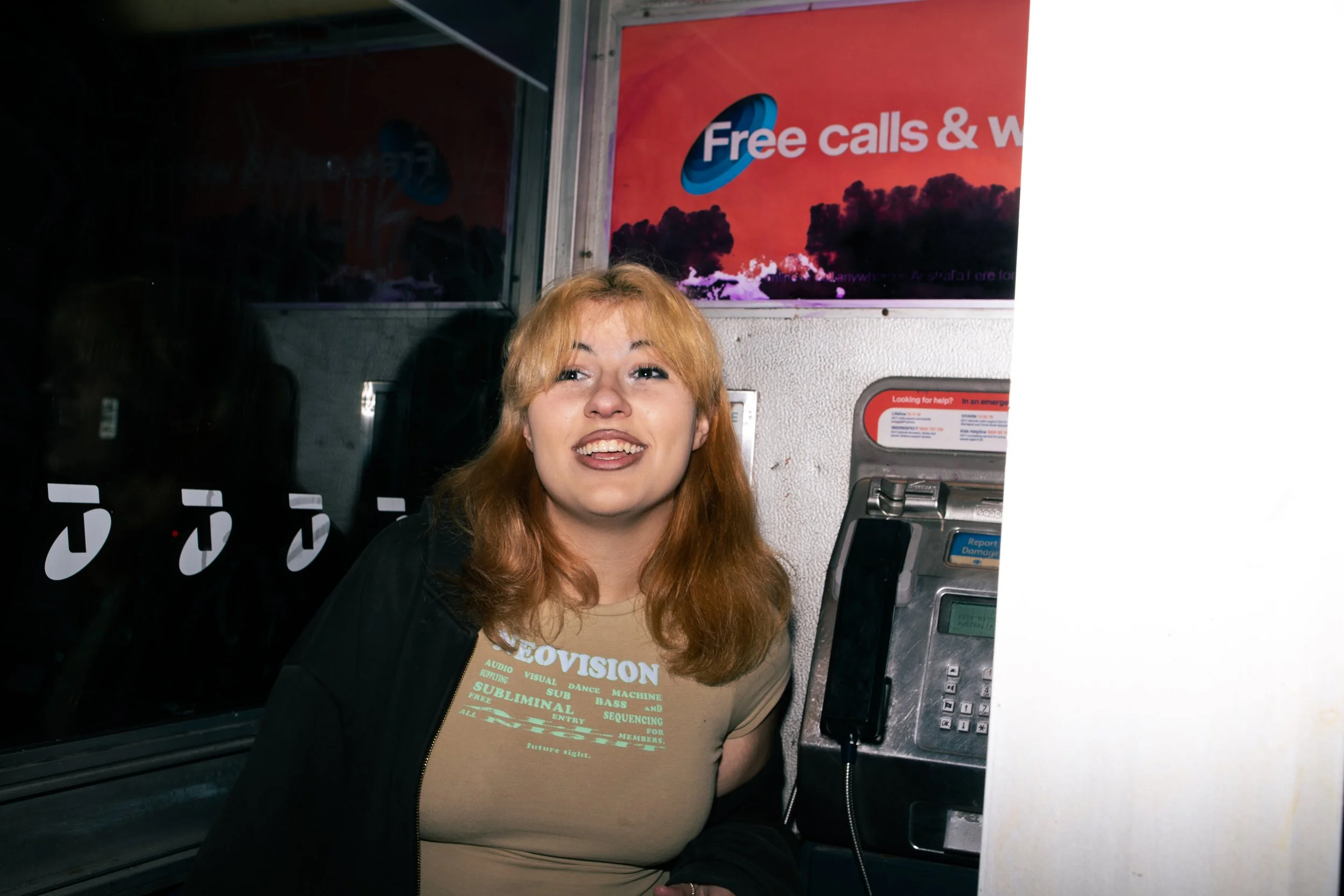 A young woman with reddish-blonde hair smiling while talking on a payphone inside a building at night.