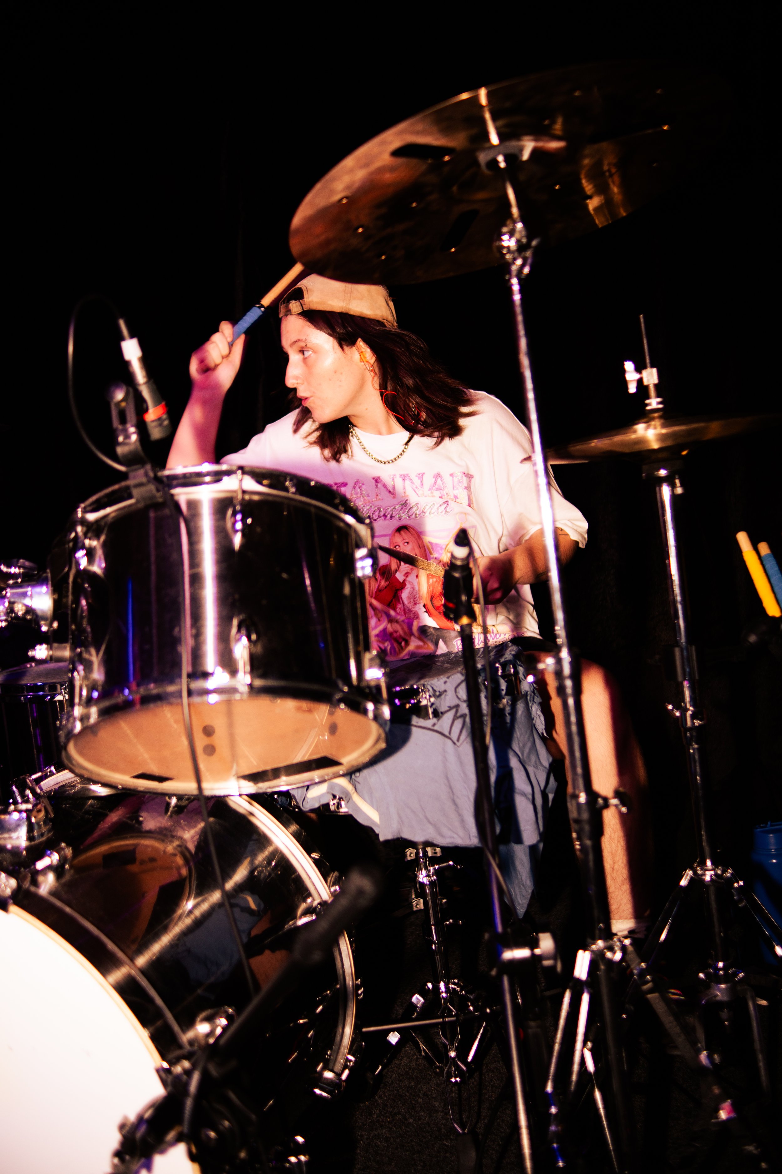 A young woman playing a drum set on stage, wearing a beige baseball cap backwards, a white graphic T-shirt, and gray shorts, with black hair and red earbuds.