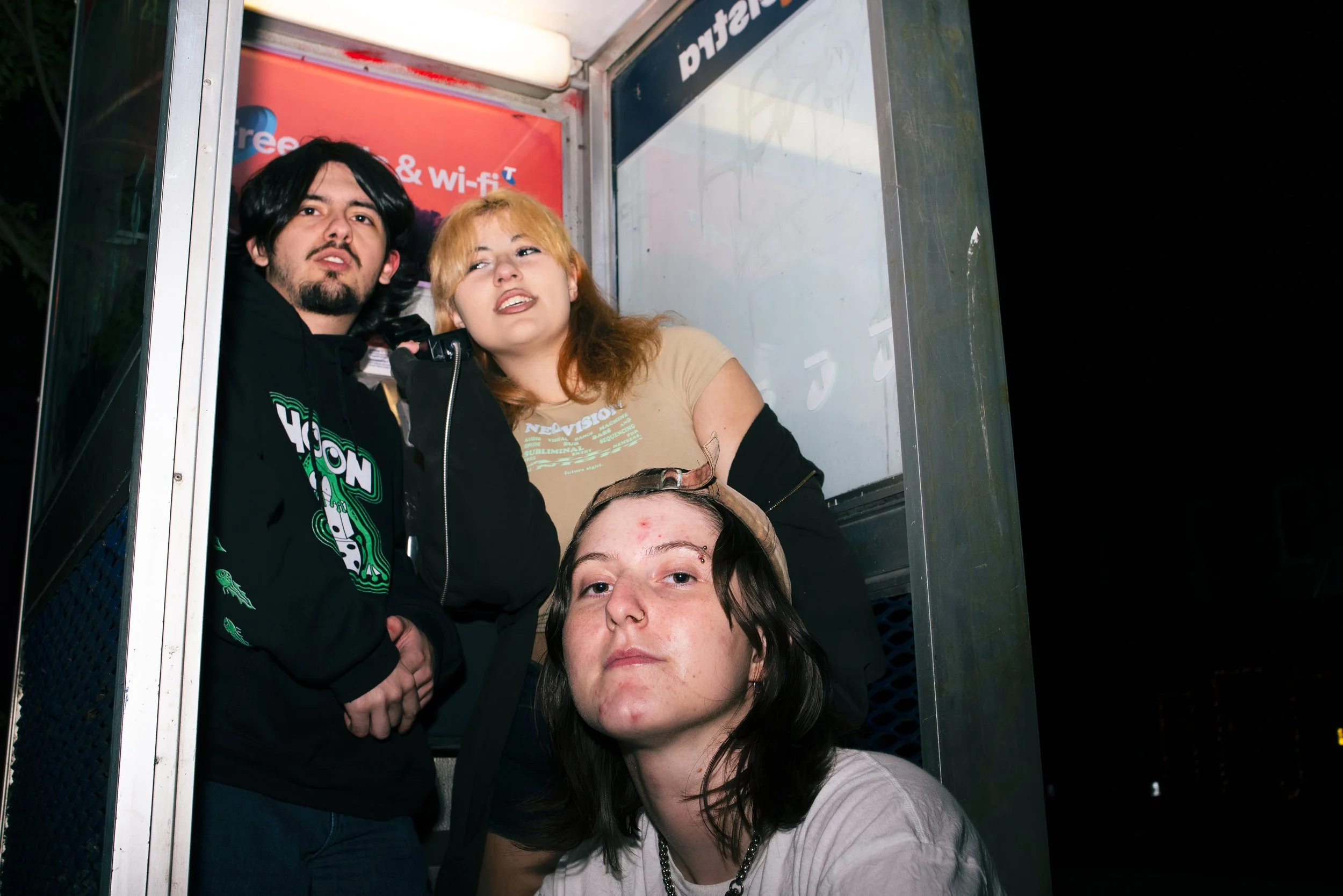 Four young people inside a phone booth at night, with posters and advertisements visible on the booth walls.