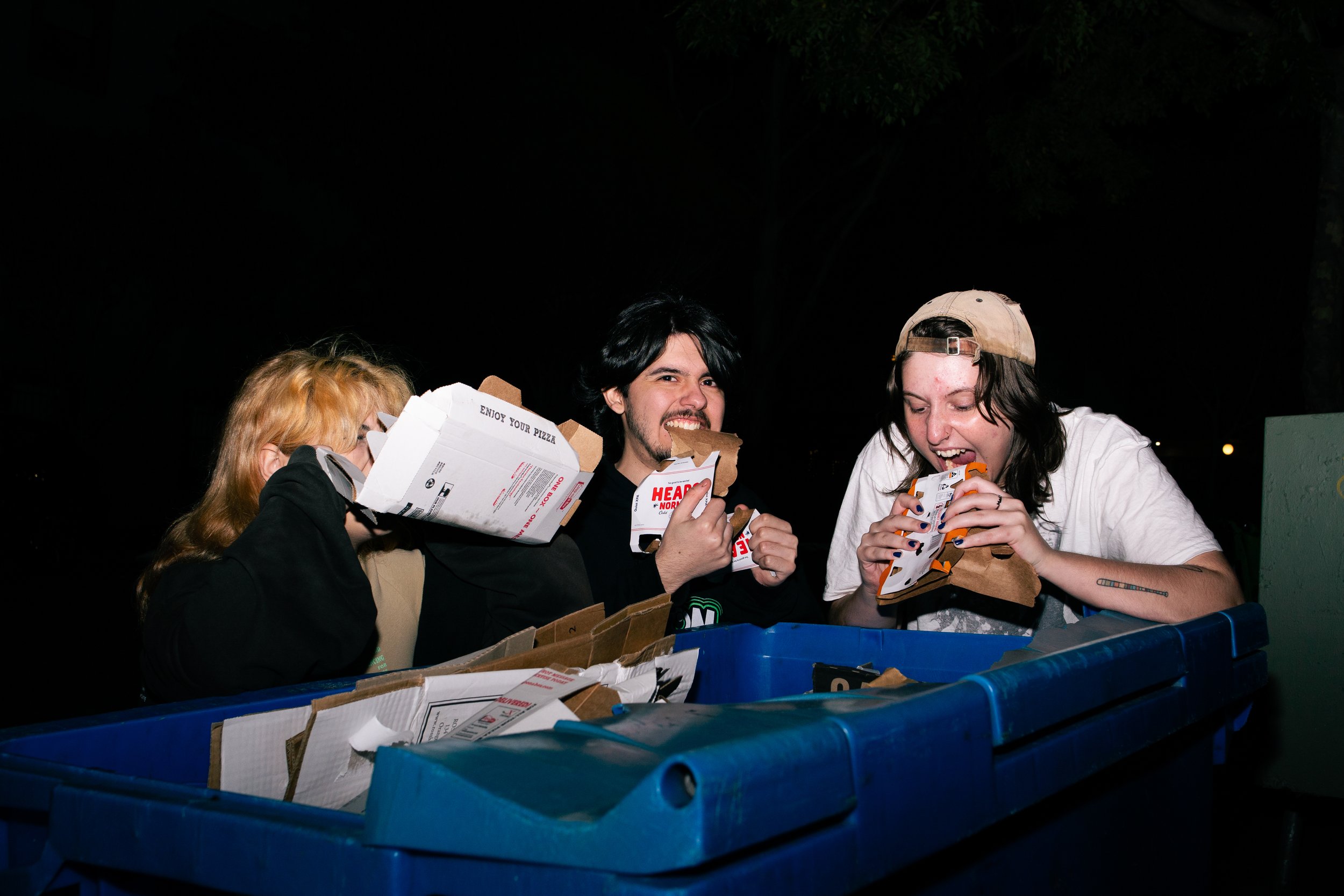 Three young people eating pizza together at night, standing behind a large blue recycling bin filled with pizza boxes.