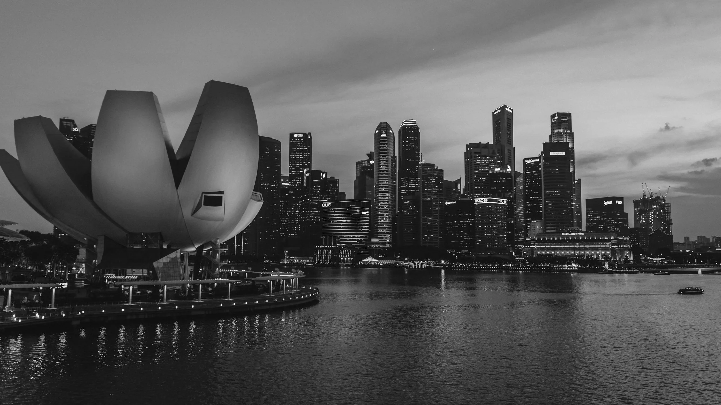 Black and white image of the Singapore skyline with the ArtScience Museum in the foreground and tall skyscrapers in the background, near the water.