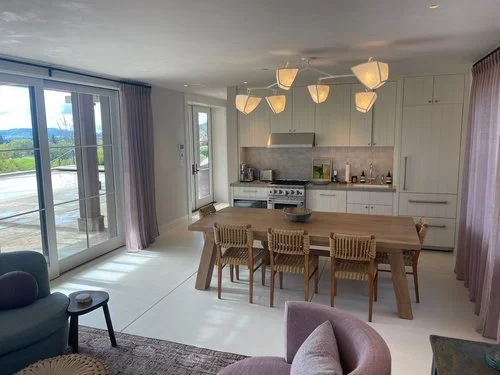 Open-concept kitchen and dining area with large windows, sliding glass door, and hanging pendant lights. The space includes a wooden dining table with six chairs, white cabinetry, and a view of a balcony and outdoor greenery.