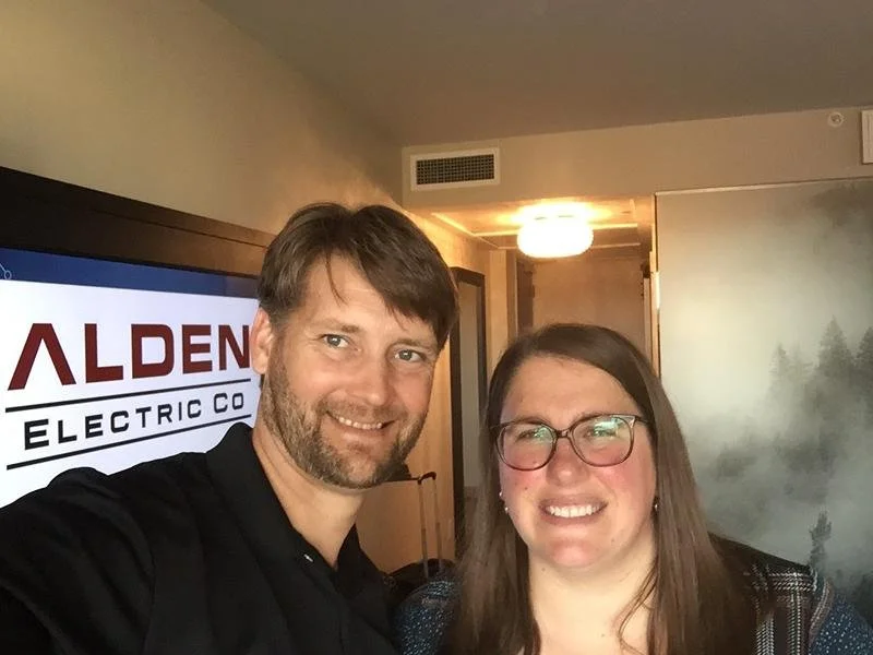 Kevin and Leah smiling for a selfie indoors with a sign that reads "ALDEN ELECTRIC CO." in the background.