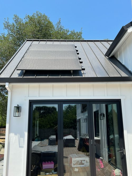 The roof of a house with a metal roof and solar panels. The house has a sliding glass door with a black frame and white exterior walls, and outside is a deck with some furniture.