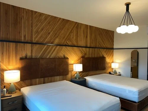 Hotel room with two beds, wooden headboards, nightstands, lamps, and a modern ceiling fixture.