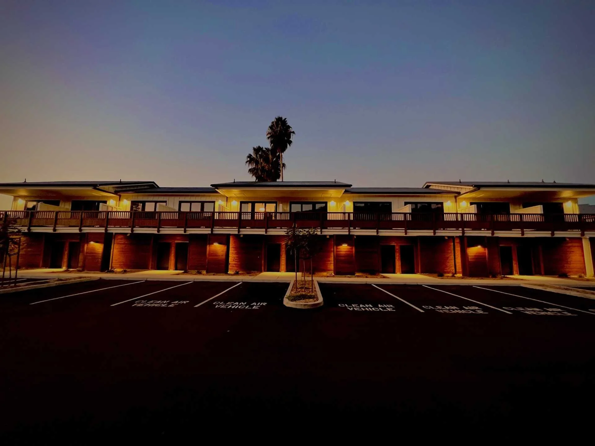 A two-story motel with external rooms, illuminated by yellow lights at dusk. There is a parking lot with marked spaces in front, labeled 'CLEAN AIR VEHICLE,' and tall palm trees in the background.