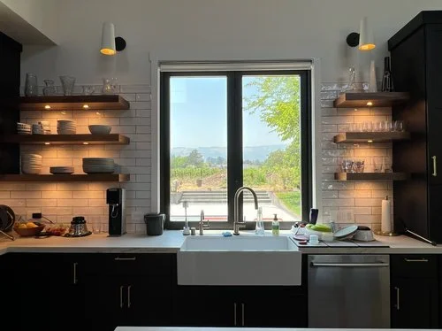 Modern kitchen with open shelving, a white farmhouse sink, and a large window overlooking a green outdoor landscape. Open shelves hold glassware and dishes, with two small wall-mounted lights illuminating the space.