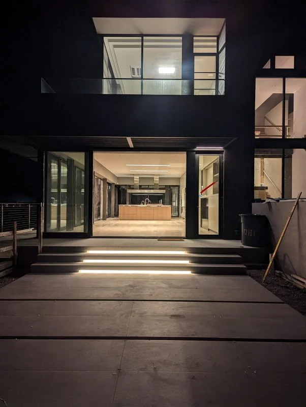 Modern two-story house at night with glass sliding doors, illuminated stairs leading to an open kitchen with a central island, and large windows on the upper floor.
