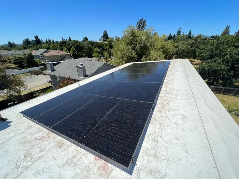 Solar panels installed on a house roof with a suburban neighborhood and trees in the background under a clear blue sky.