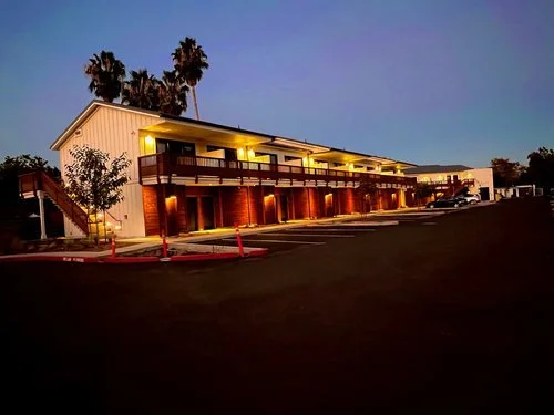 Two-story motel with exterior corridor, balconies, and palm trees, illuminated at dusk.