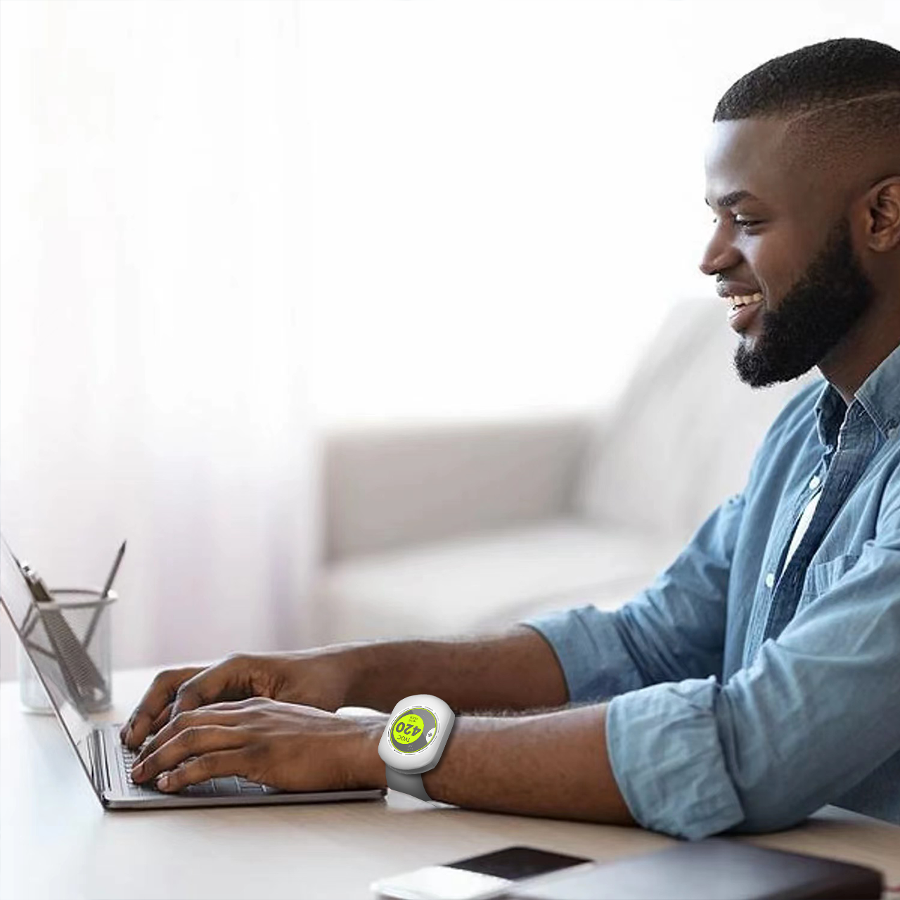 A man with a beard sitting at a desk, smiling, using a laptop, wearing a smartwatch on his left wrist with a green screen showing 420.