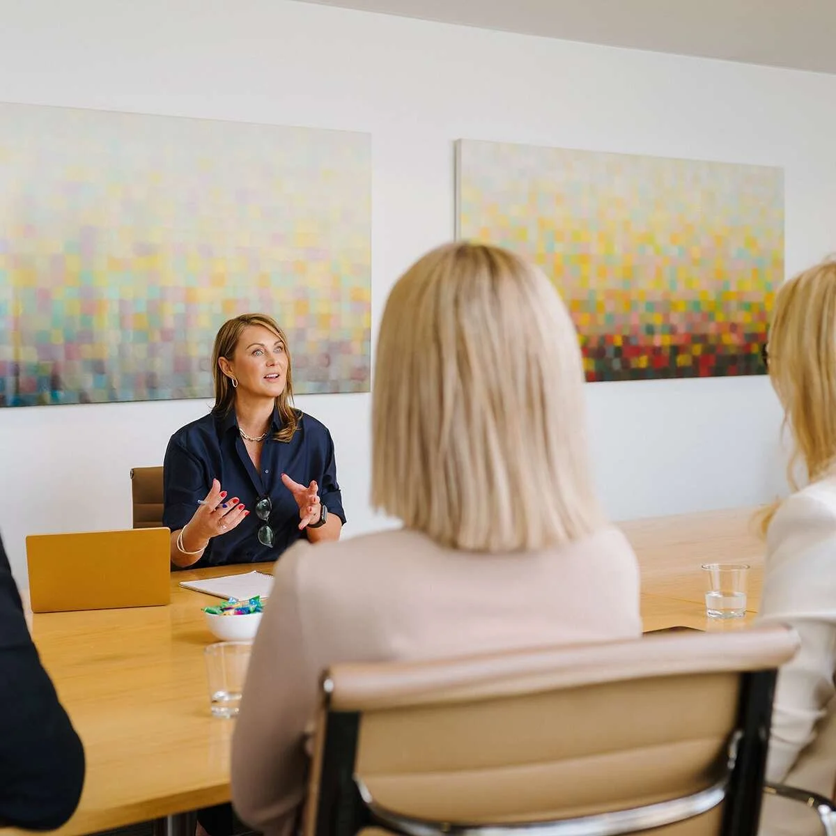 A woman in a navy shirt leading a meeting with three women sitting around a wooden table, two with blonde hair and one with brown hair. The woman speaking is gesturing with her hands, and there are abstract artwork on the wall behind her.
