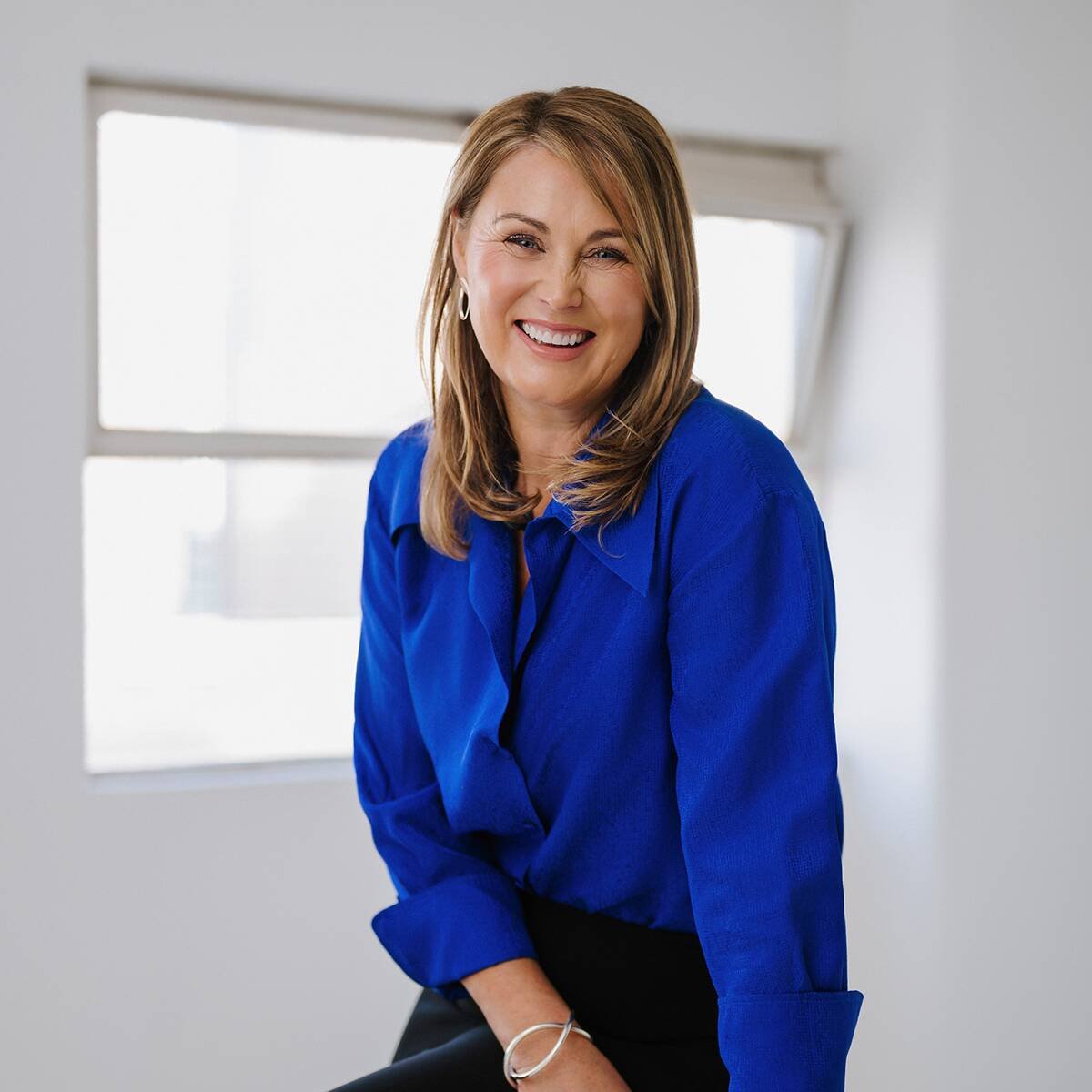 A woman with shoulder-length light brown hair, wearing a bright blue blouse, smiling and sitting in a brightly lit room with a window in the background.