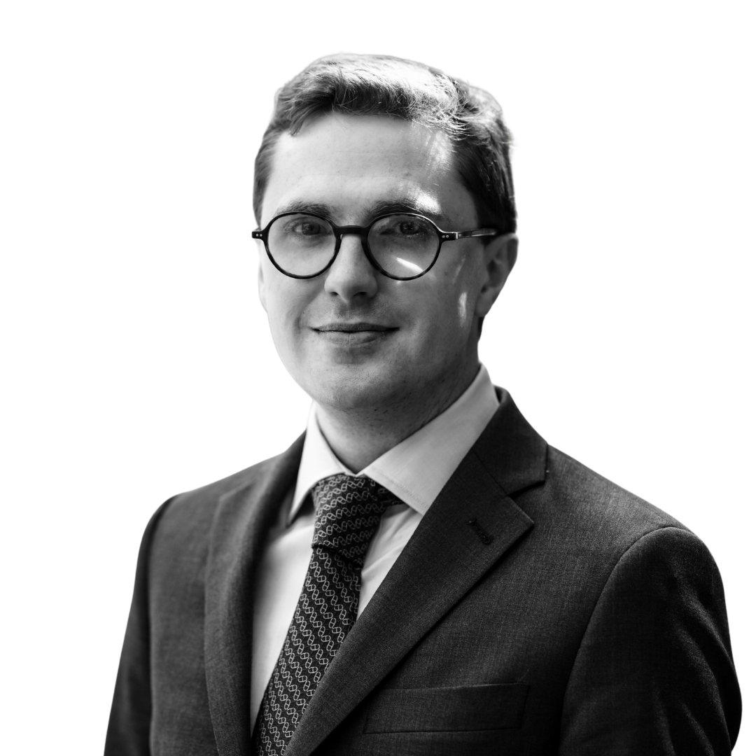 Black and white portrait of Dean W. Ball wearing glasses, a suit, and a patterned tie, smiling, against a plain background.