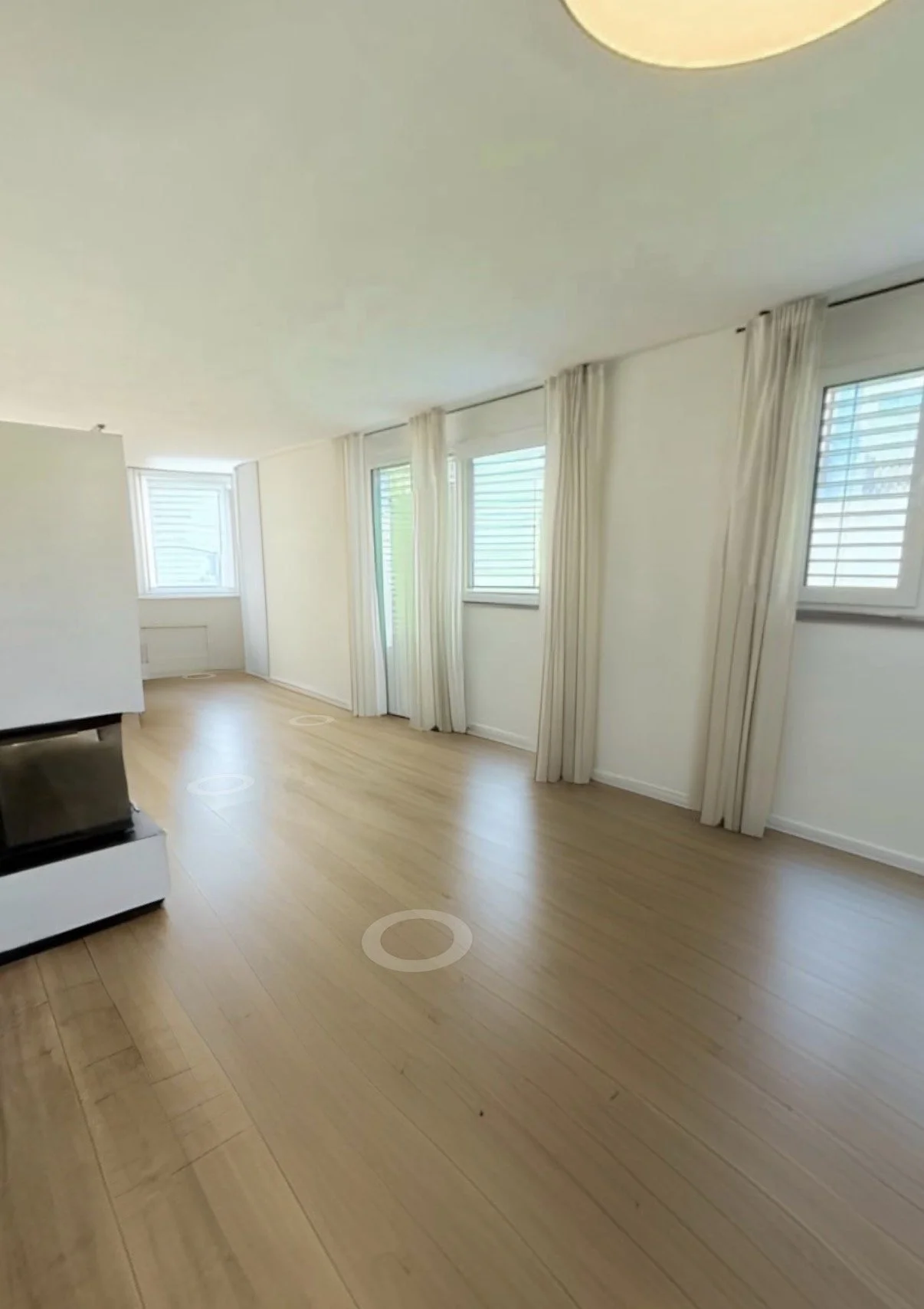 Ein leeres Wohnzimmer mit hellen Wänden, Holzboden, mehreren Fenstern mit weißen Jalousien und weißen Vorhängen.