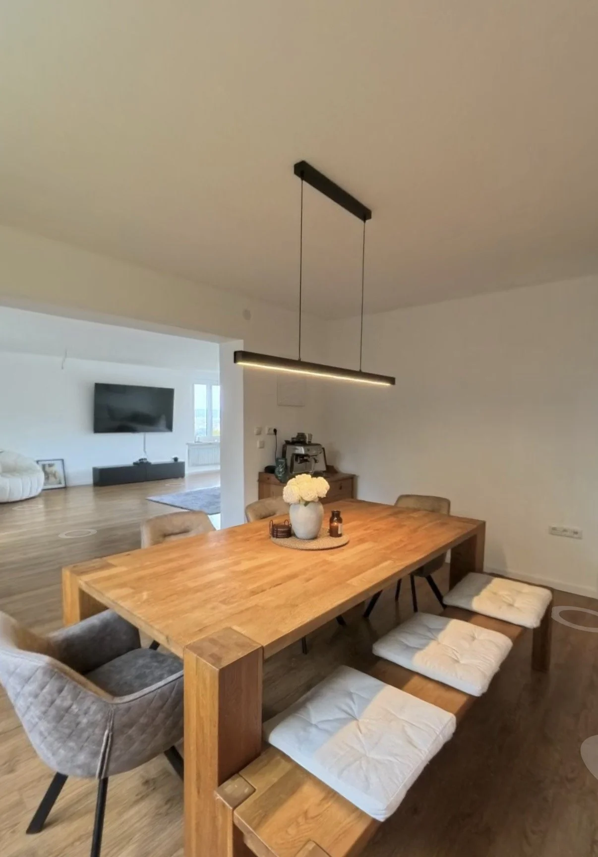 Ein moderner Essbereich mit einem großen Holztisch, Sitzkissen und Stühlen, einer weißen Vase mit Blumen, und einer modernen Lampe über dem Tisch.