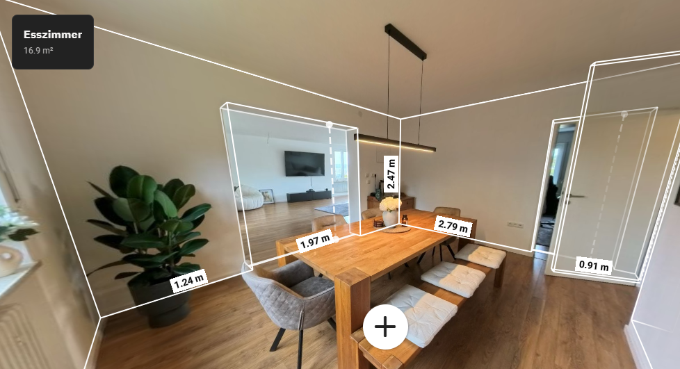 Ein stilvoller Essraum mit einem großen Holztisch, Sitzkissen auf einer Bank, einem bequemen Sessel, einer großen Zimmerpflanze und einer TV-Webenseite im Hintergrund.