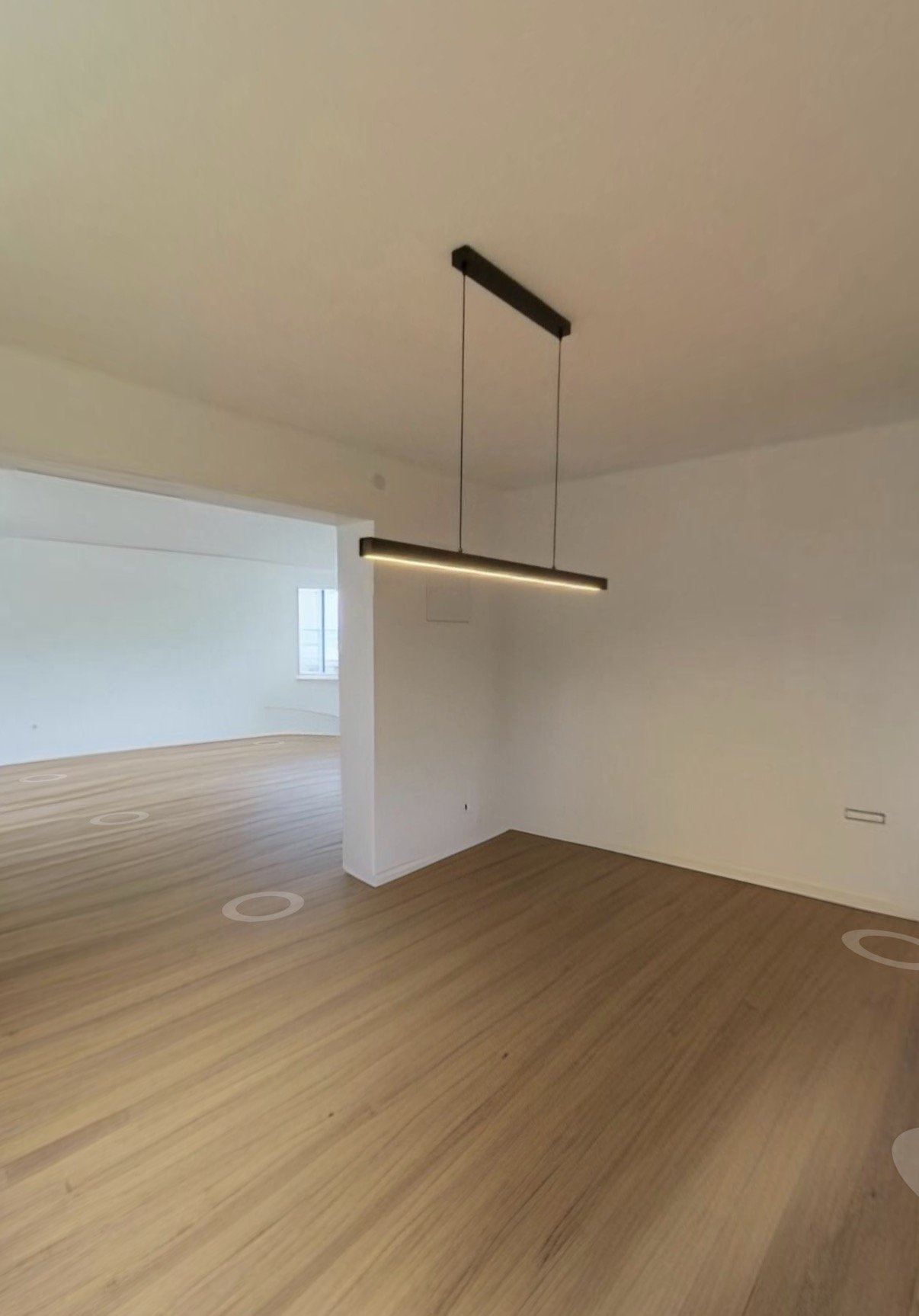 Helles, modernes Wohnzimmer mit Holzboden, weiß verfegelten Wänden und einer schwarzen, langen Deckenleuchte.