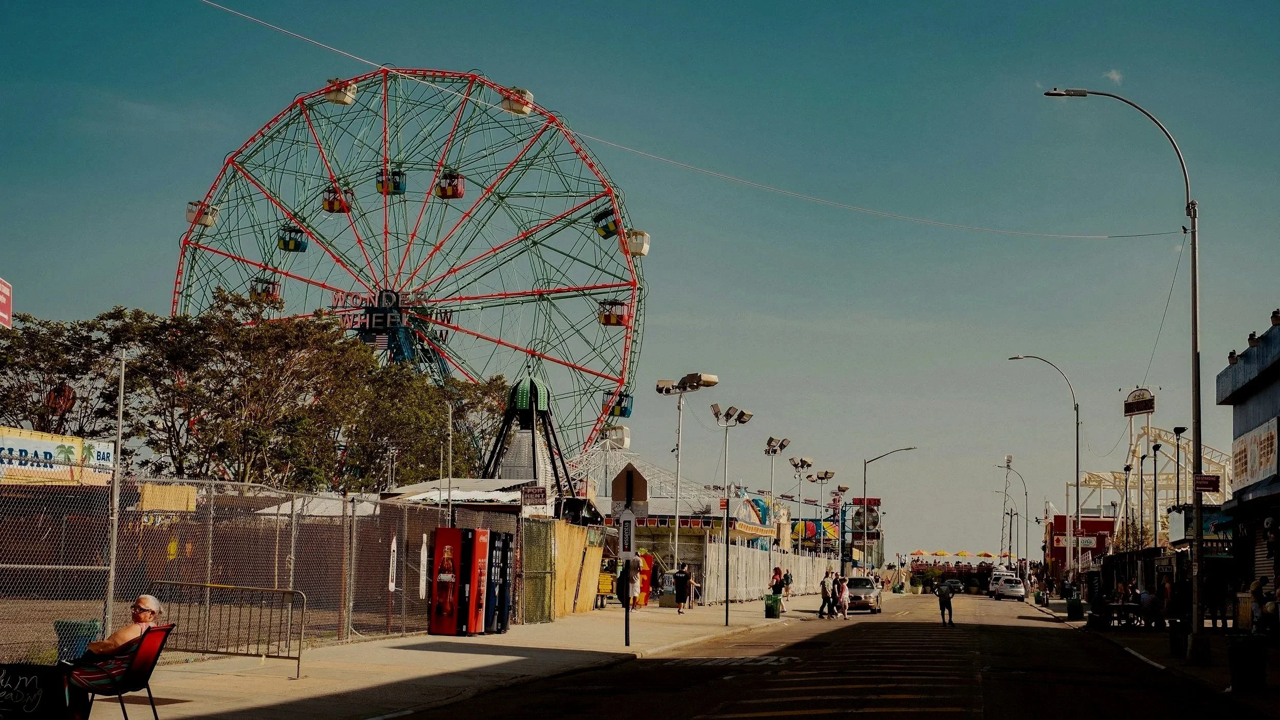 Sonnenuntergang auf einem Volksfest mit Riesenrad, Menschen auf der Straße, Stände und Karussells im Hintergrund.