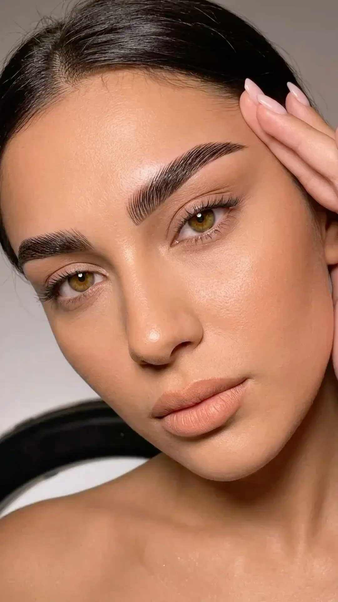 Close-up of a woman with dark hair, well-groomed eyebrows, and natural makeup, touching her temple with her hand.