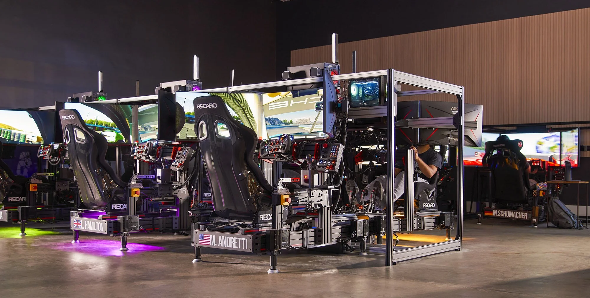 People using racing simulators with large screens and realistic setups. The simulators have Recaro seats and various controls, and users are wearing headphones and gloves while engaging in a virtual racing experience.